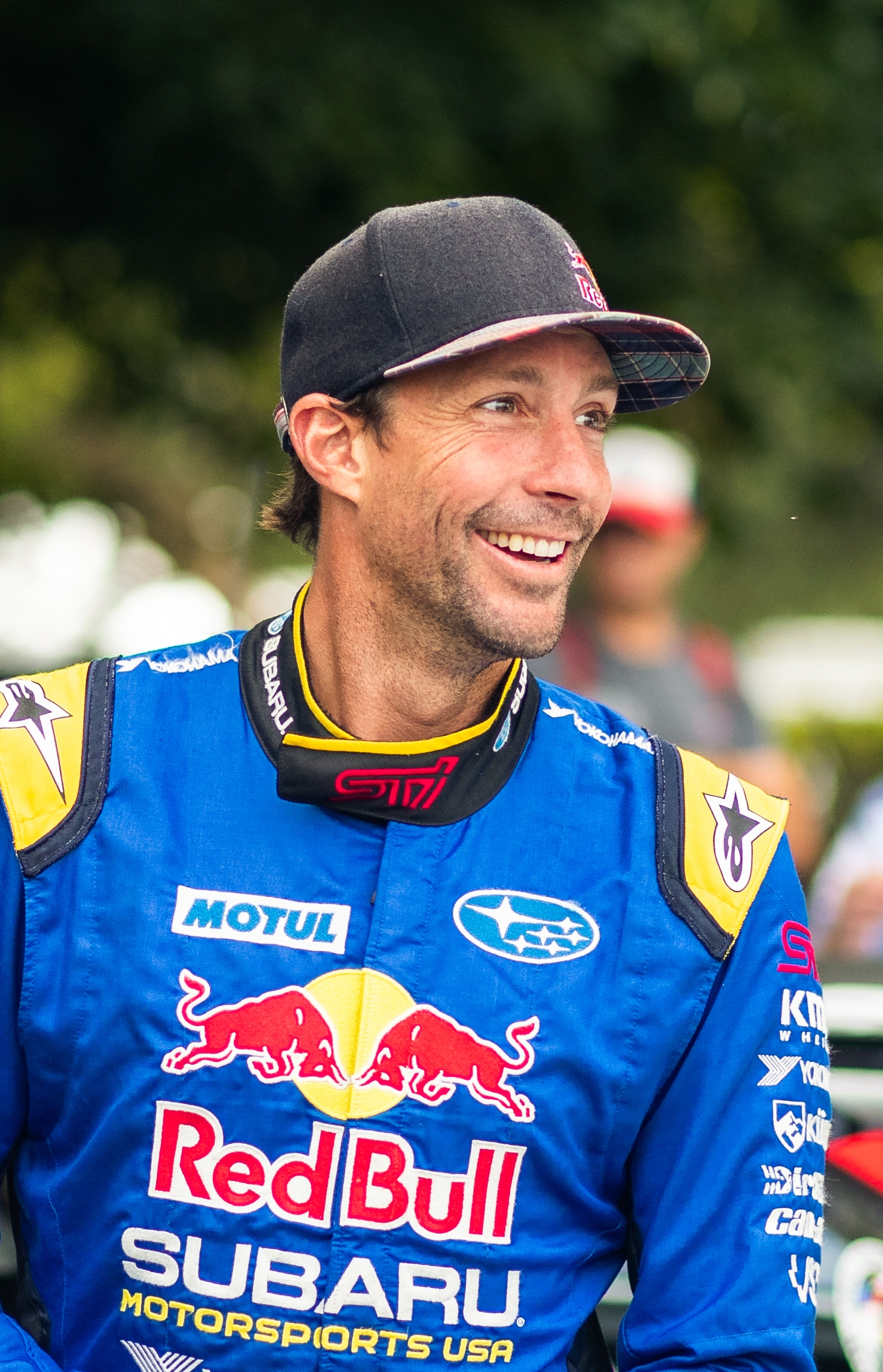 Travis Pastrana in a blue Red Bull Subaru racing suit stands next to a black and white race car.