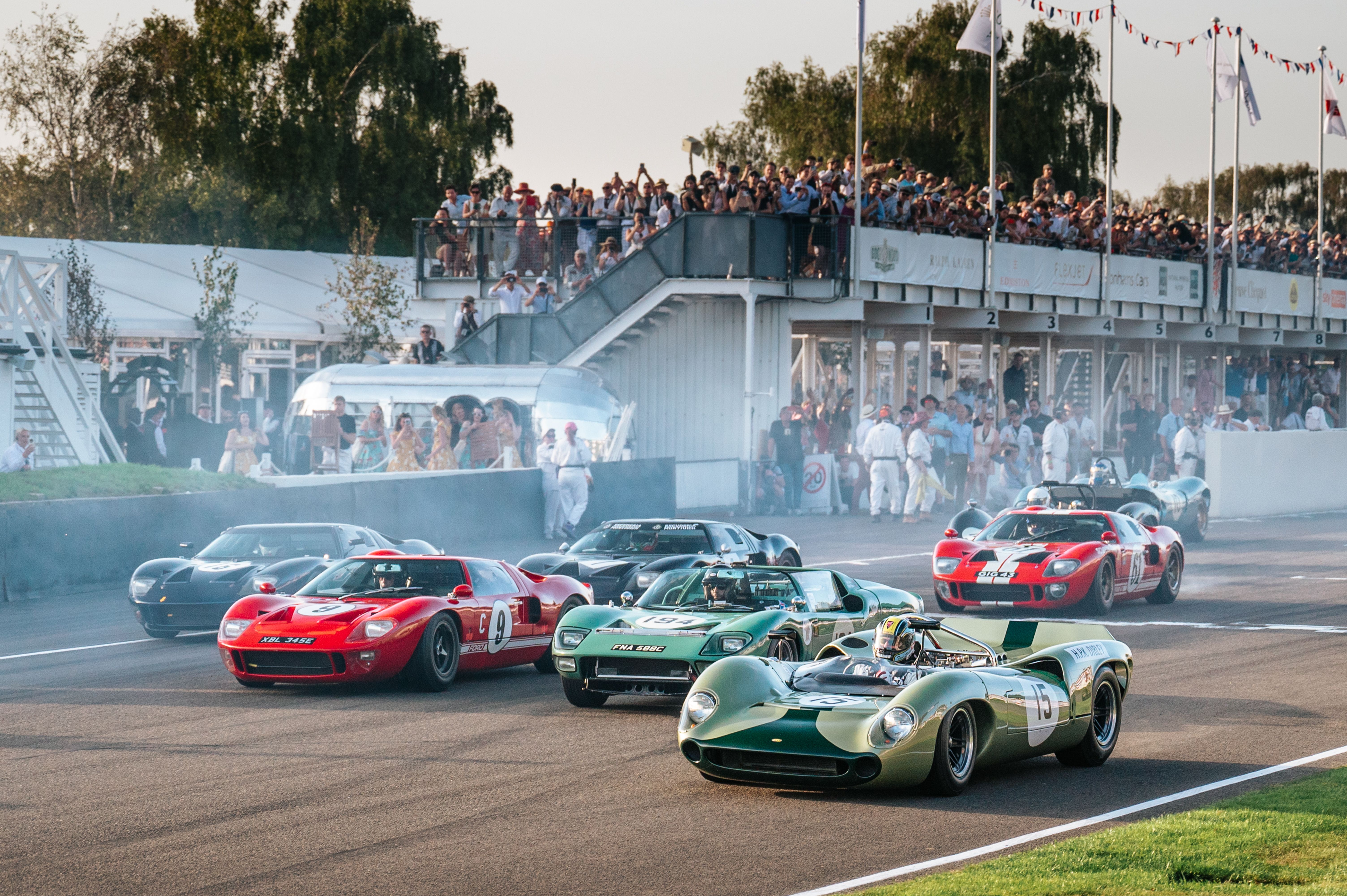 Classic race cars on a track, kicking up smoke, with spectators in a grandstand.