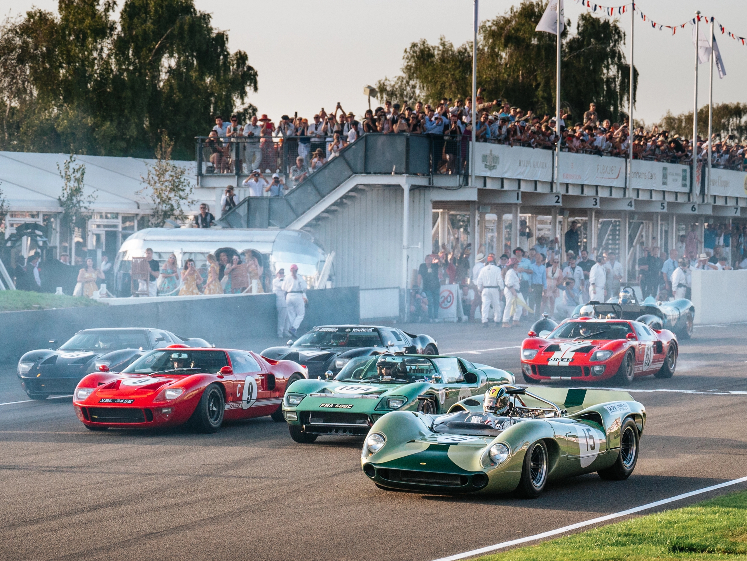 Classic race cars on a track, kicking up smoke, with spectators in a grandstand.