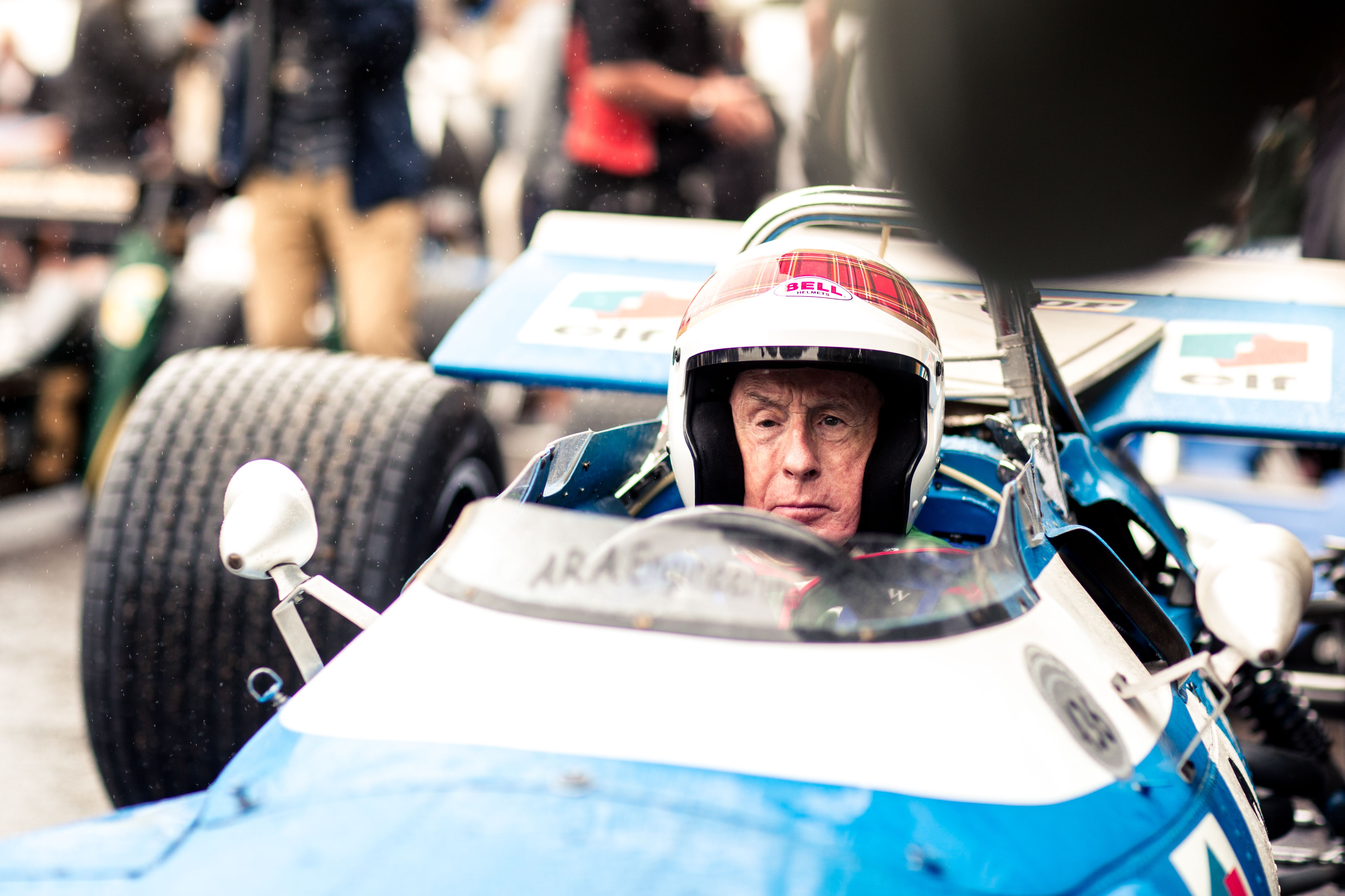 Sir Jackie Stewart, wearing a tartan helmet, sits in a vintage blue and white Formula 1 car.