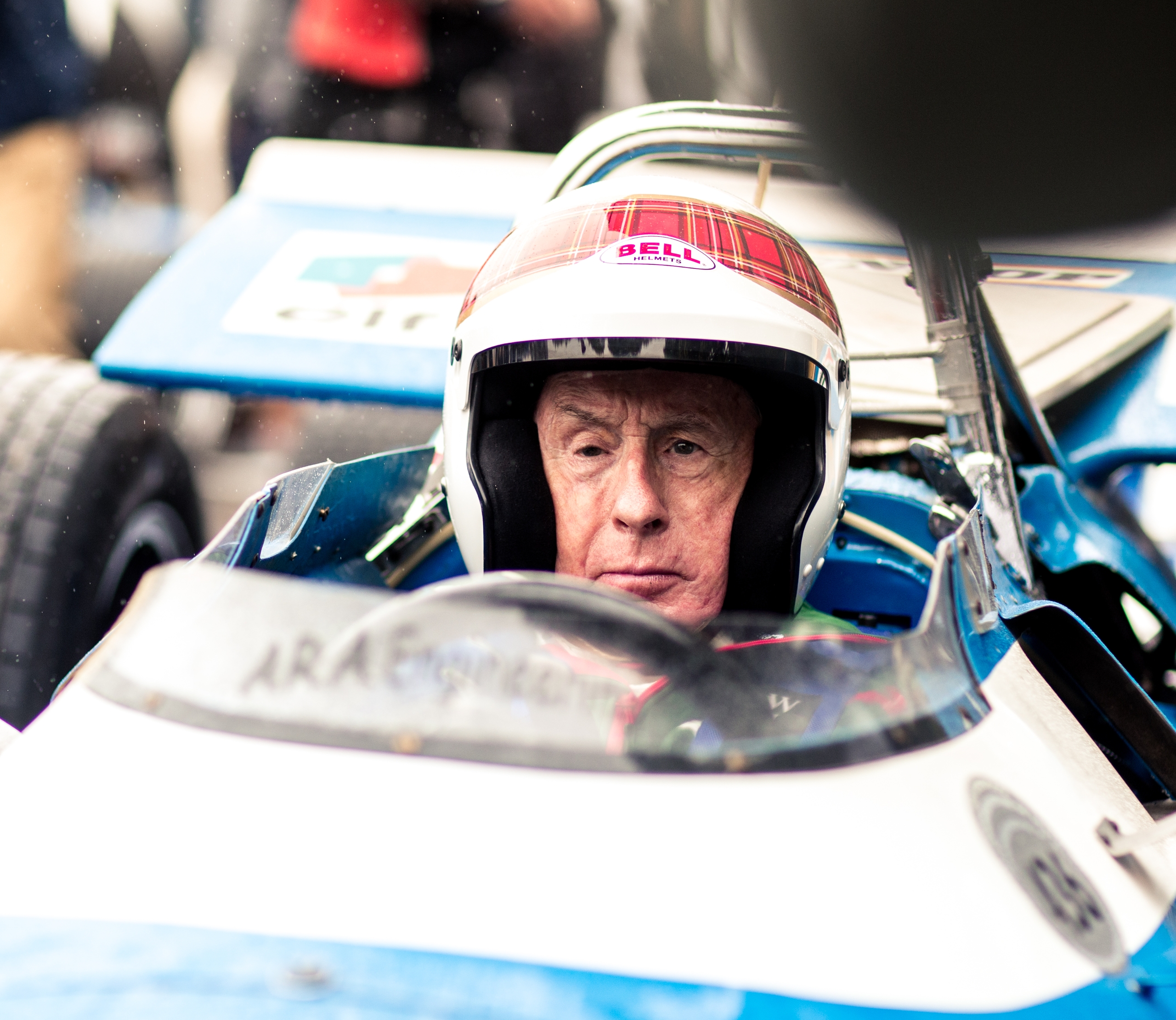 Sir Jackie Stewart, wearing a tartan helmet, sits in a vintage blue and white Formula 1 car.