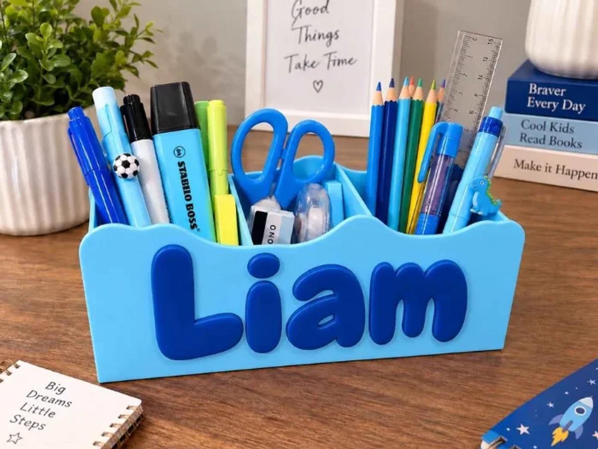 A blue 3D printed desk organiser with a name in raised letters across the front, holding pens, pencils and scissors.