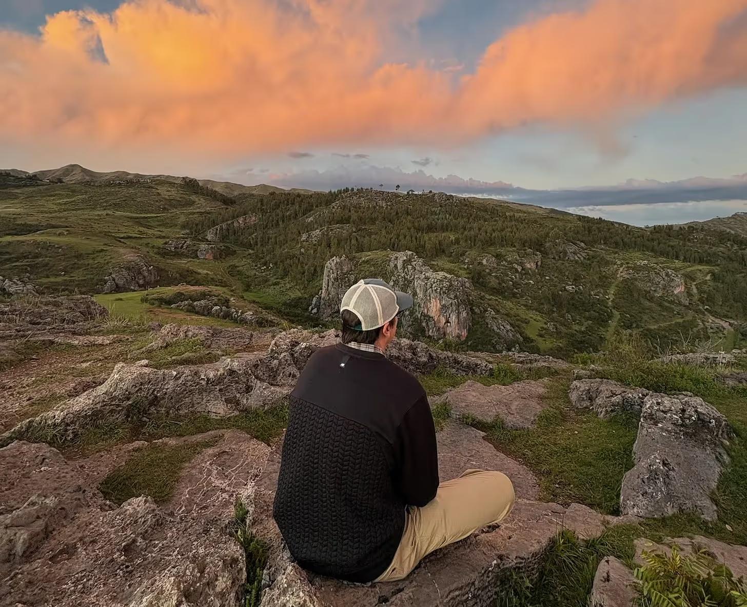 Man sitting on a rocky hilltop looking out over green hills at sunset