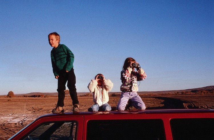 Three kids standing on the roof of a red car, two looking through binoculars at a desert landscape