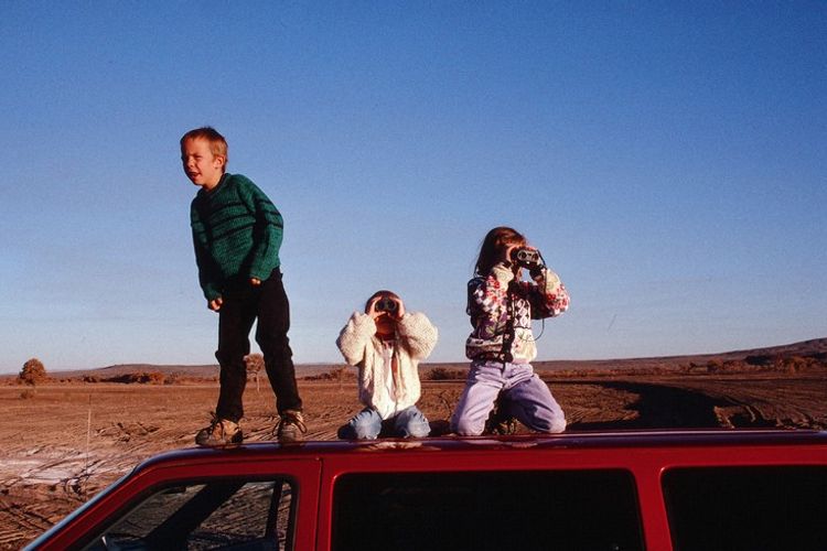 Three kids standing on the roof of a red car, two looking through binoculars at a desert landscape