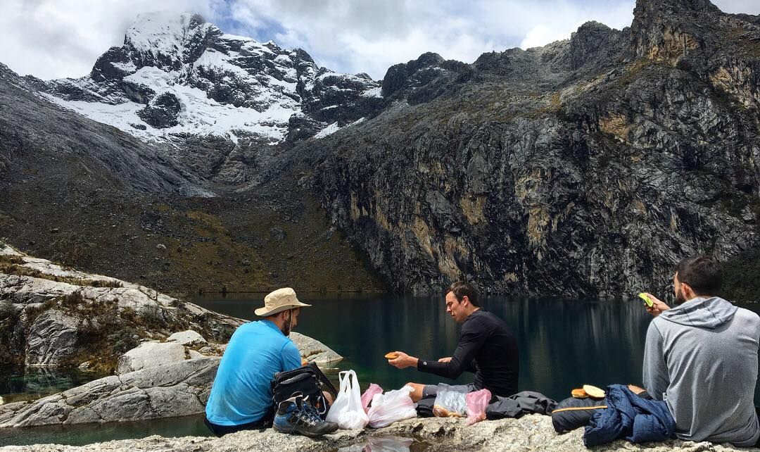Three hikers resting by an alpine lake beneath snow-capped peaks