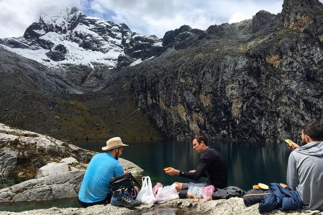 Three hikers resting by an alpine lake beneath snow-capped peaks