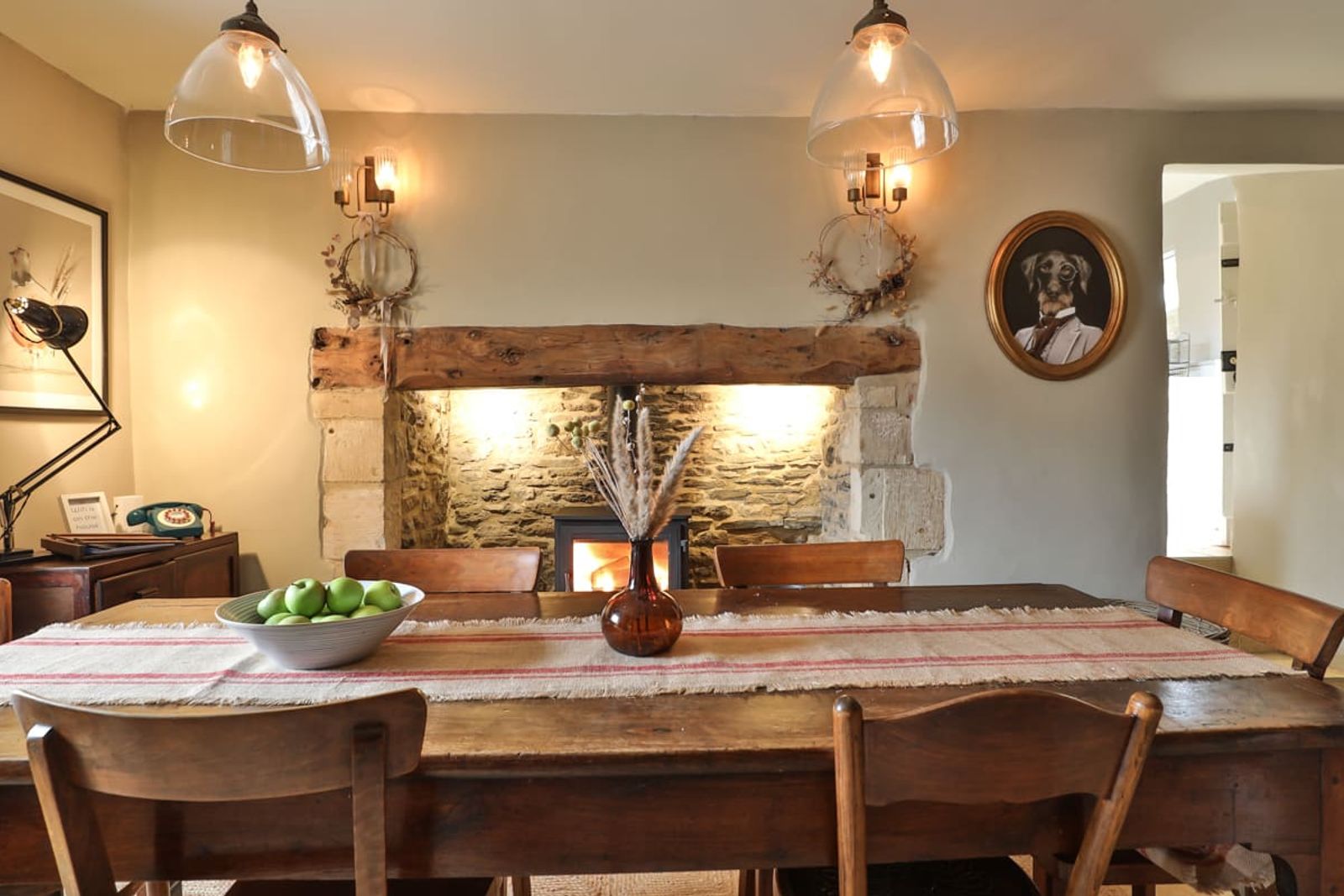 Cottage dining room with a farmhouse table under   twin glass pendants, lit wood-burning stove in a stone   fireplace, and a framed dog portrait on the wall.
