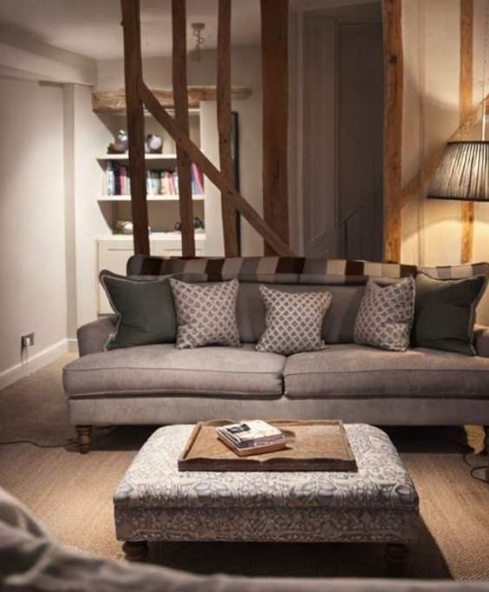Cottage sitting room with exposed diagonal structural beams  overhead, a grey sofa layered with green and patterned  cushions and a striped throw, a built-in bookshelf to the  left, and a patterned ottoman with a wooden tray of books in  front.