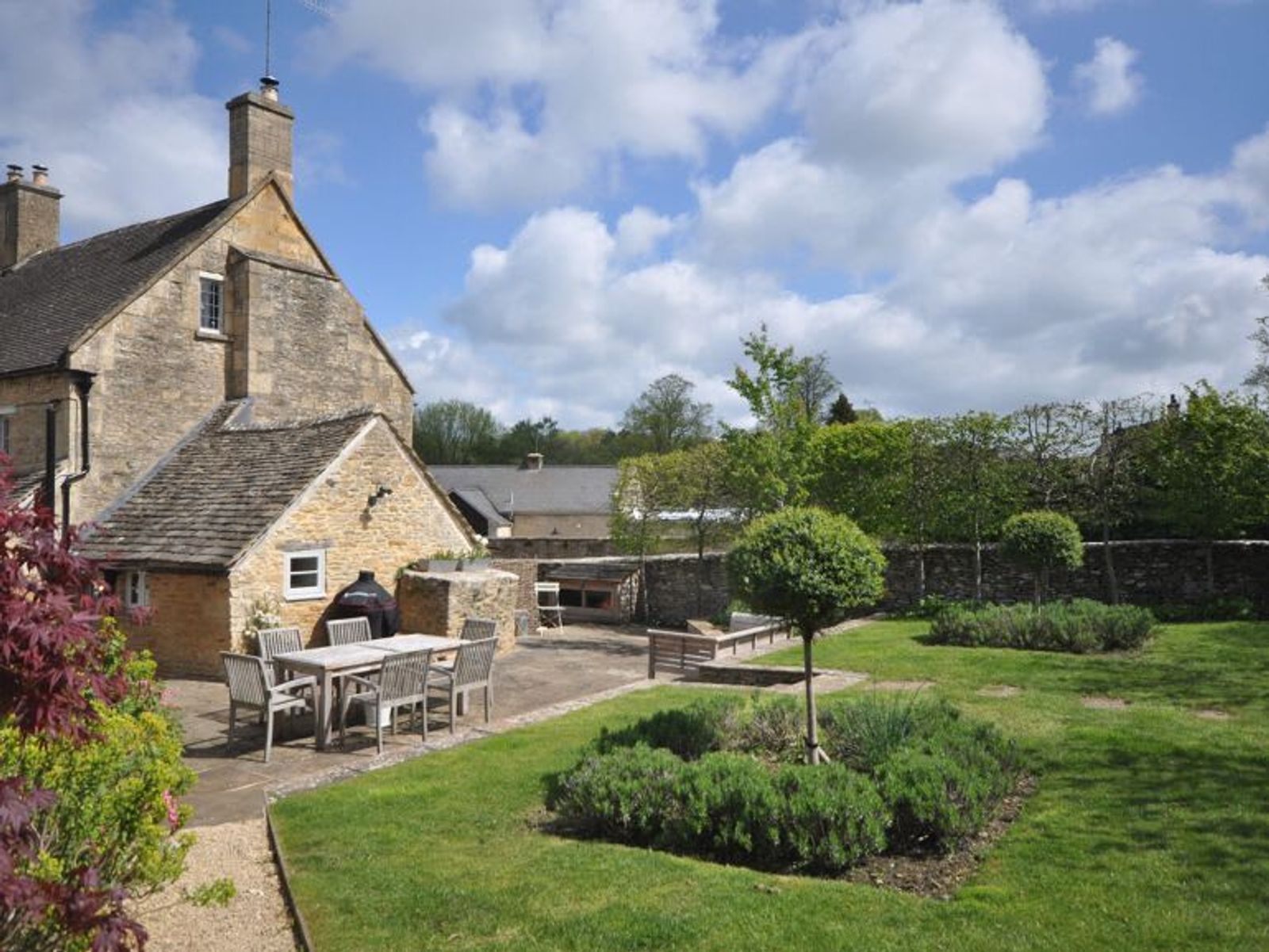 Honey-stone Cotswold cottage with a walled garden, an outdoor dining table set for six on the patio, formal lavender beds and topiary, and village rooftops beyond the garden wall under a blue sky
