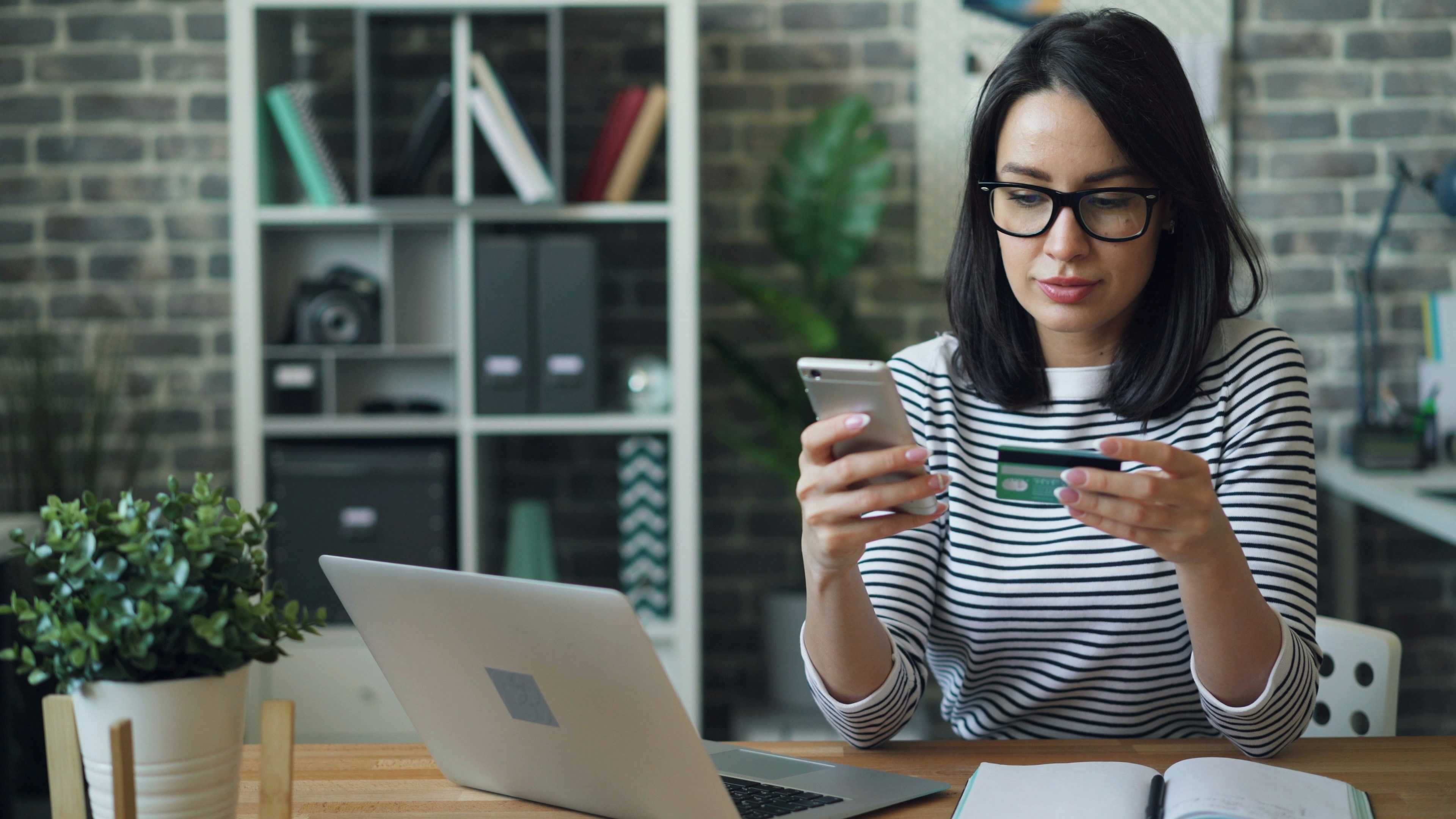 Woman sitting at desk with a laptop, holding a smartphone and credit card for online shopping.