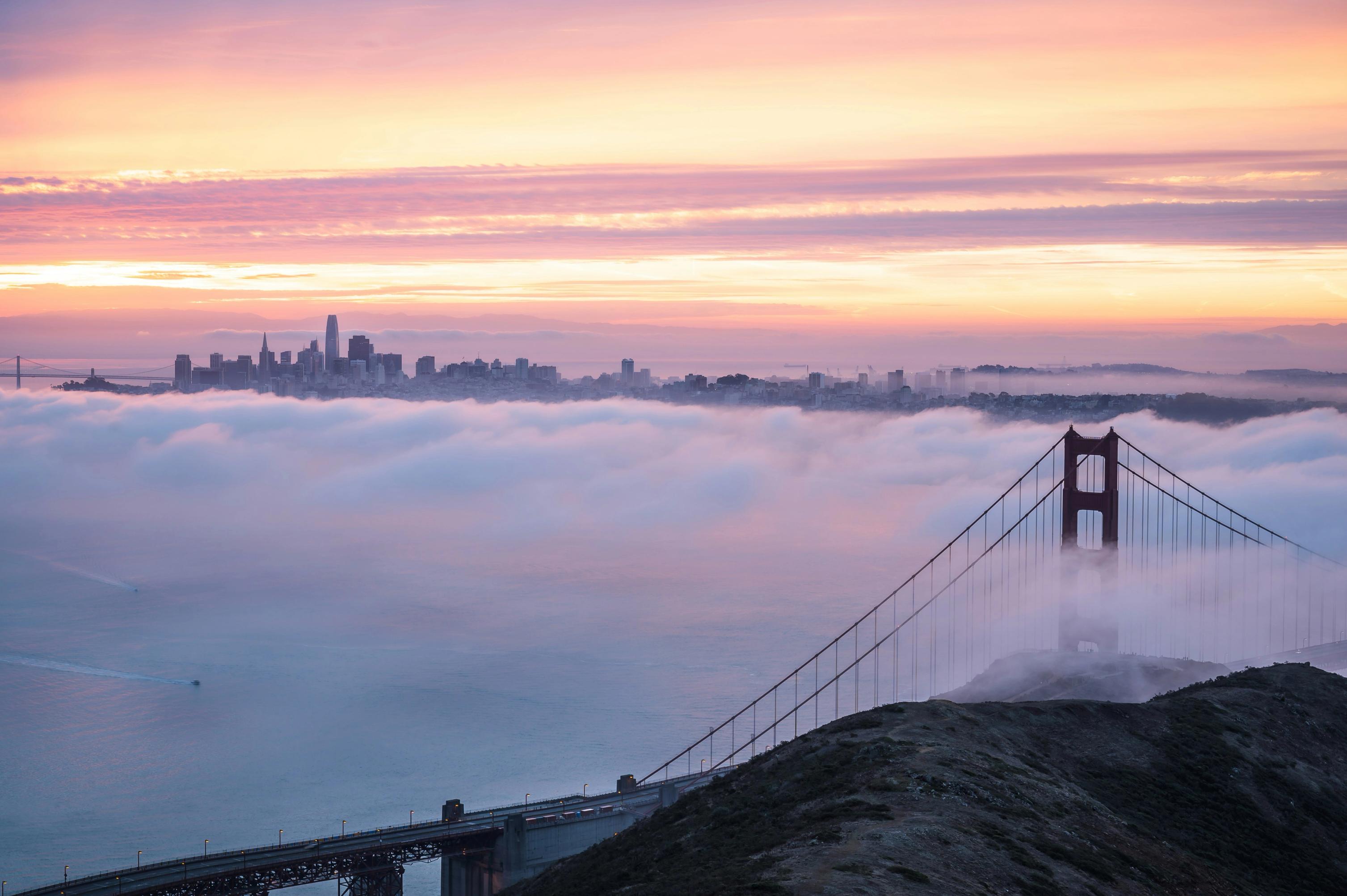 Golden Gate Bridge emerging through morning fog with San Francisco skyline in the background at sunrise.