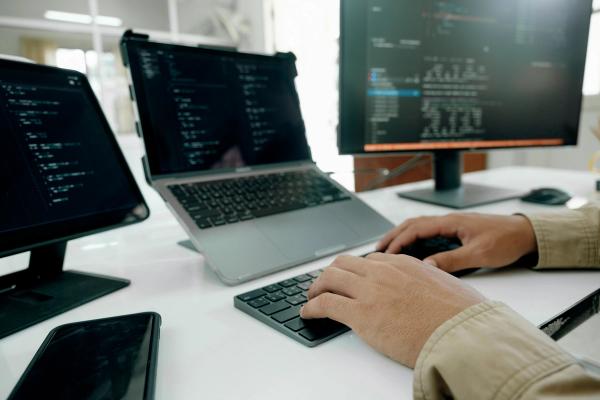 Person working on a laptop with multiple monitors displaying code, representing UI/UX design implementation and testing.