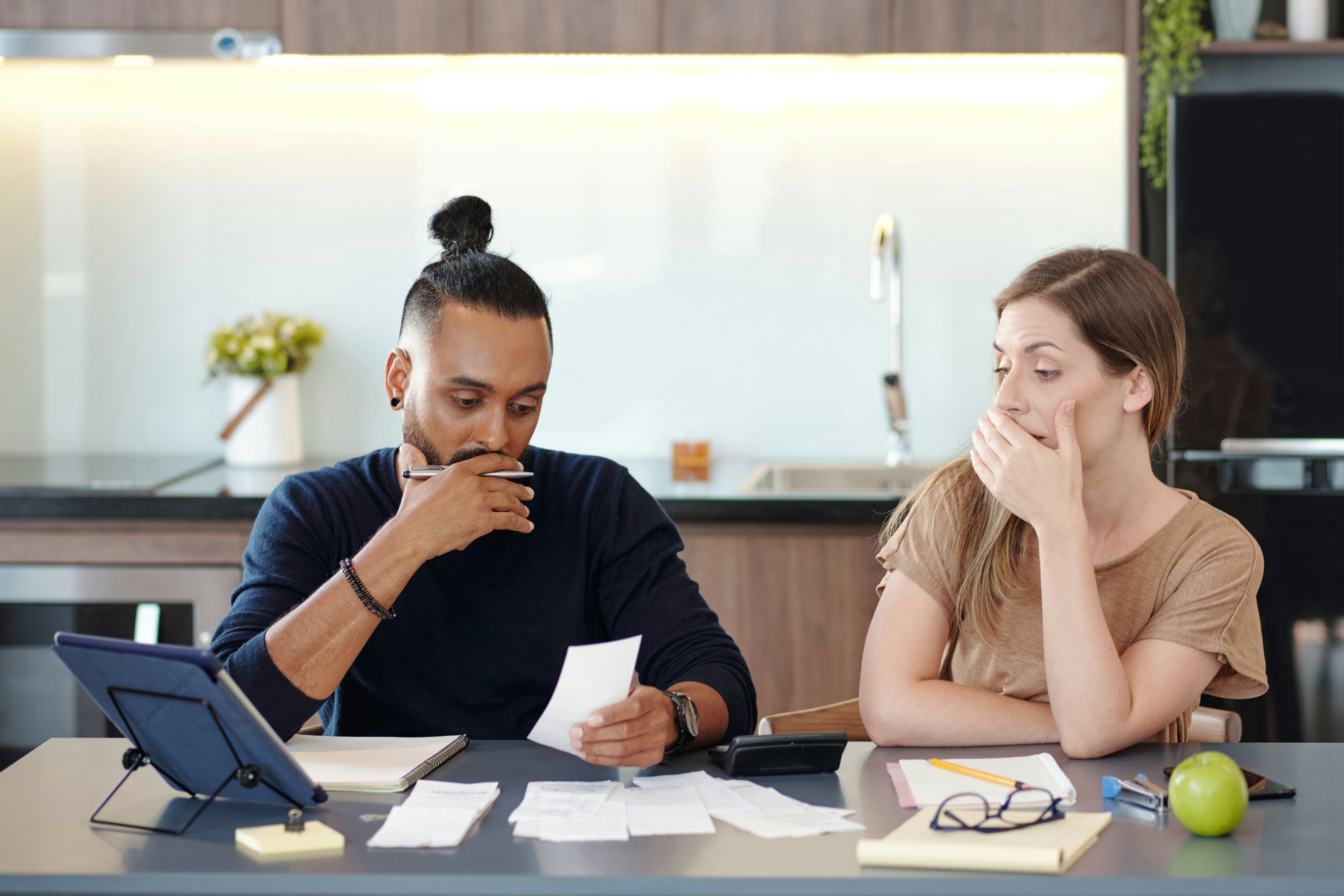 A man and a woman sitting at a kitchen table looking stressed while reviewing financial receipts and a tablet, symbolizing the overwhelming nature of "Decision Debt" and unresolved paperwork.