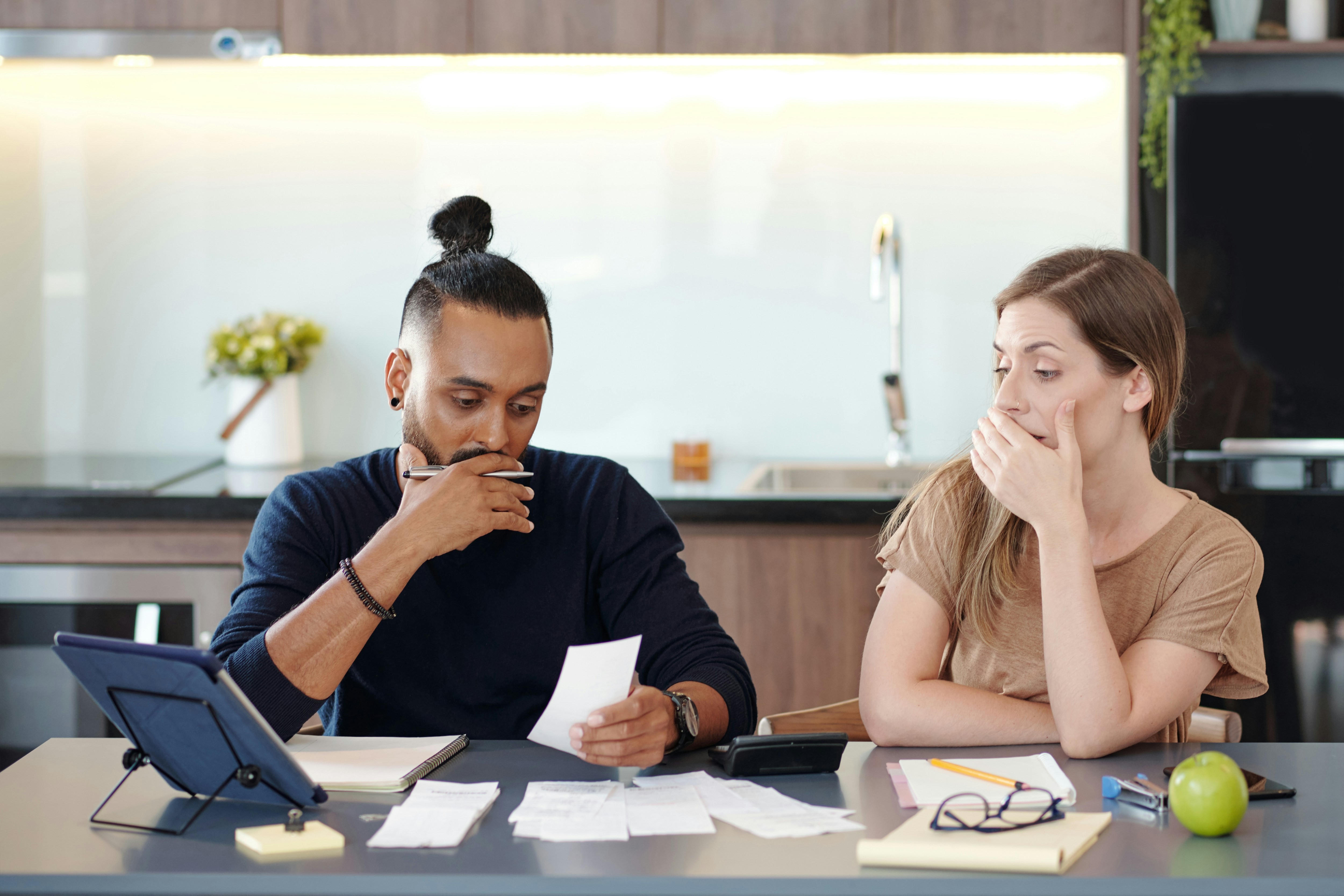 A man and a woman sitting at a kitchen table looking stressed while reviewing financial receipts and a tablet, symbolizing the overwhelming nature of "Decision Debt" and unresolved paperwork.