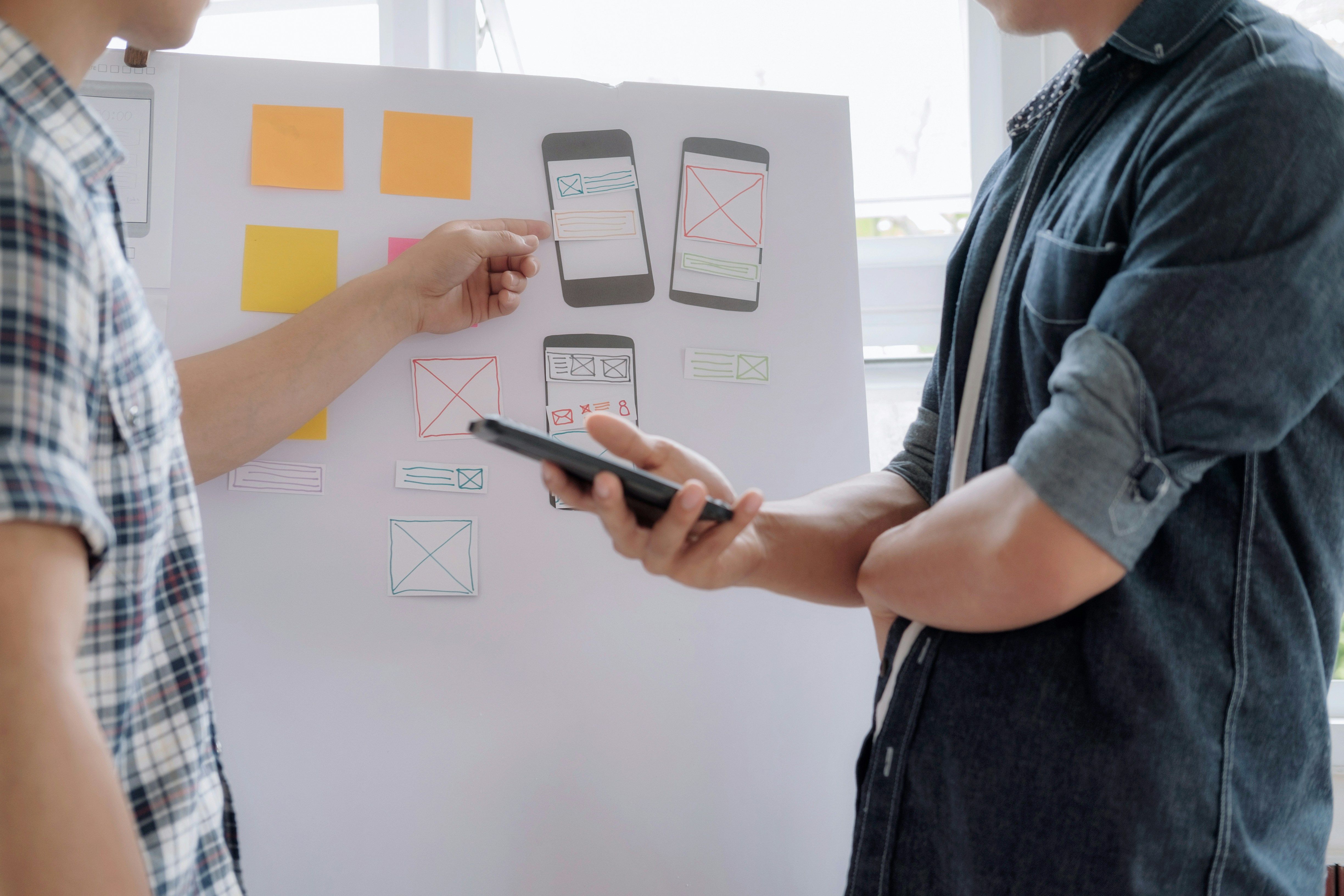 Two designers reviewing mobile app wireframes on a whiteboard during a UI/UX design planning session.