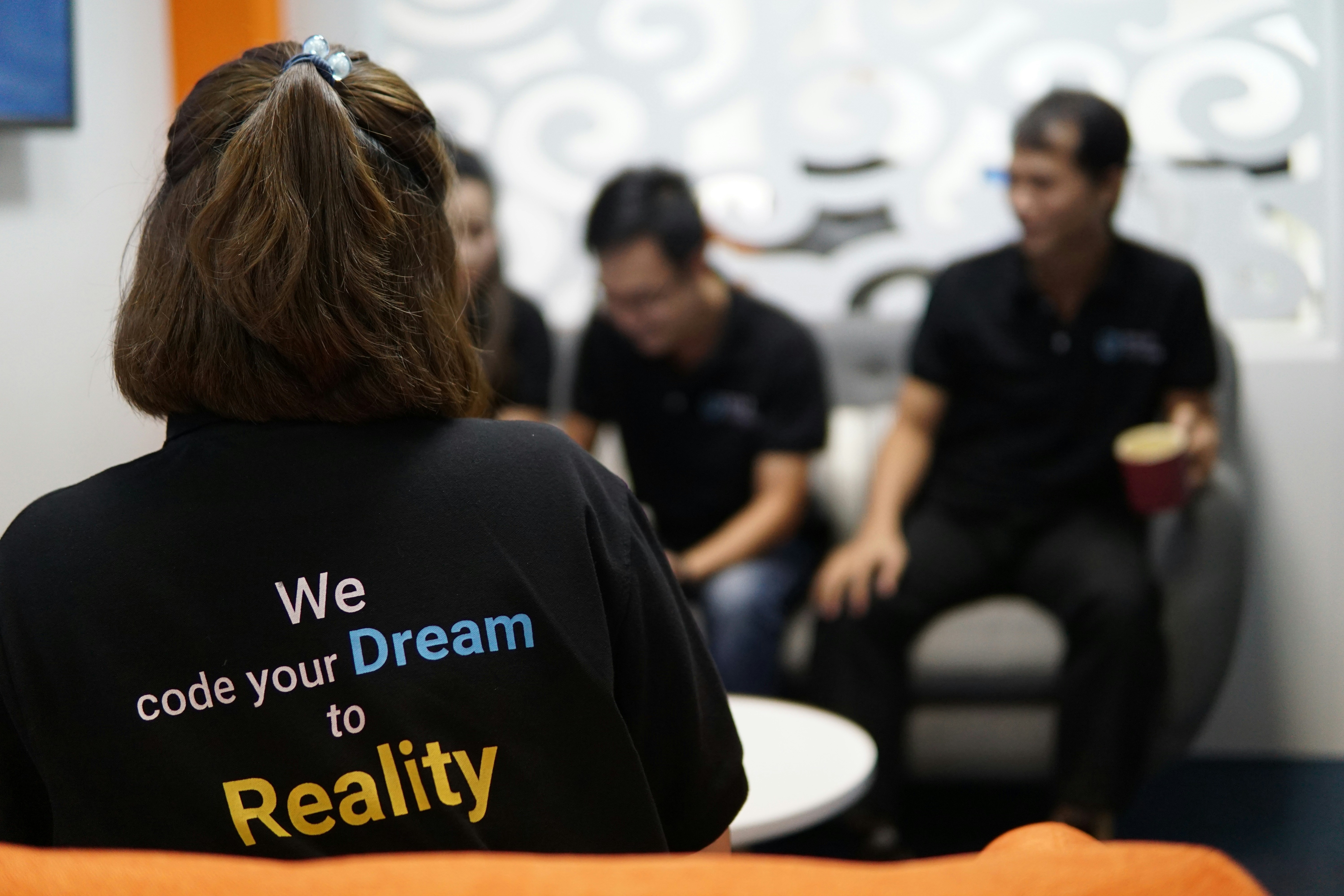 Person wearing a shirt with the text ‘We code your Dream to Reality’ sitting with a group of colleagues.