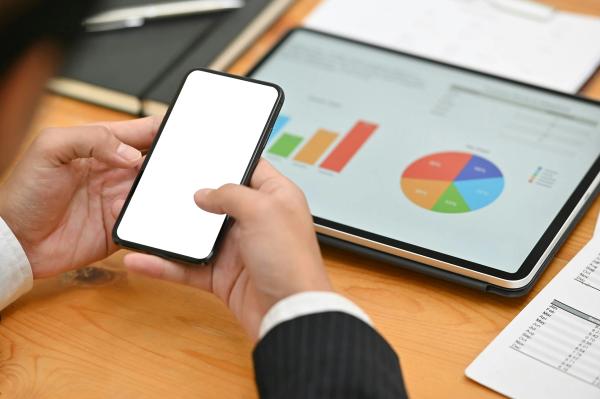 Person holding a smartphone with a tablet displaying charts and graphs for data analysis.