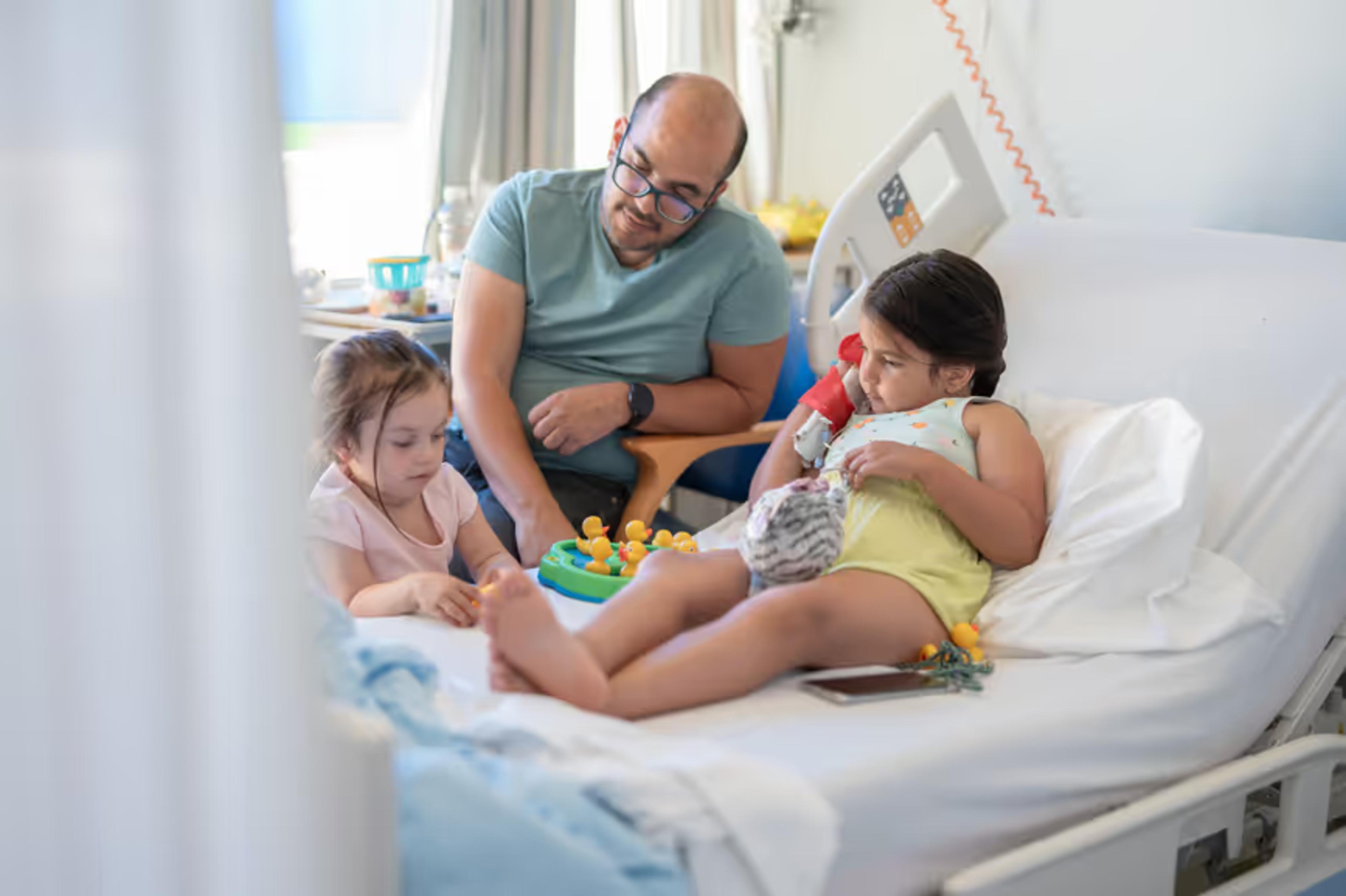 A father sits with two young children on a hospital bed