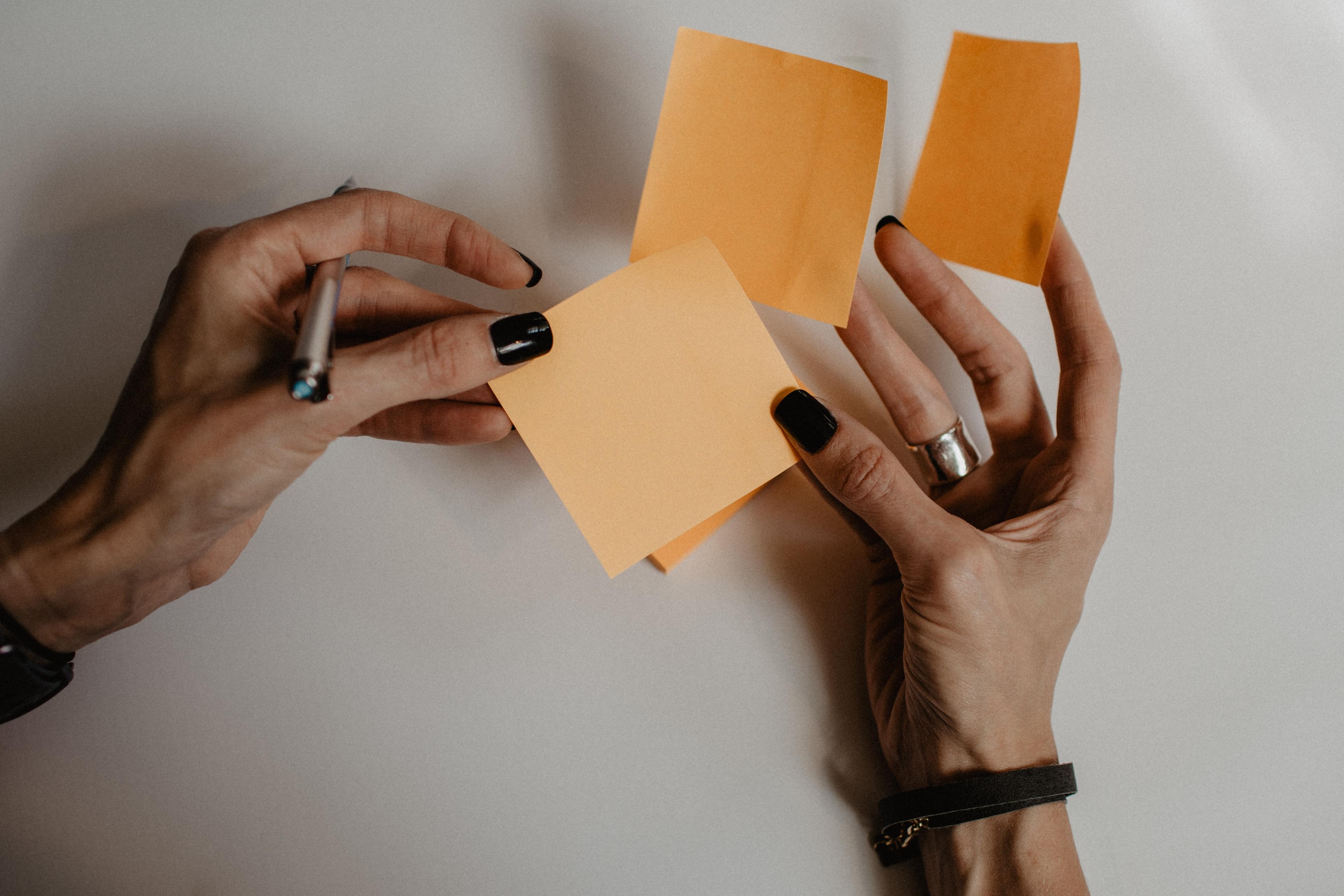Orange post-it notes (blank) in the hands of a lady with black nails holding a pen.