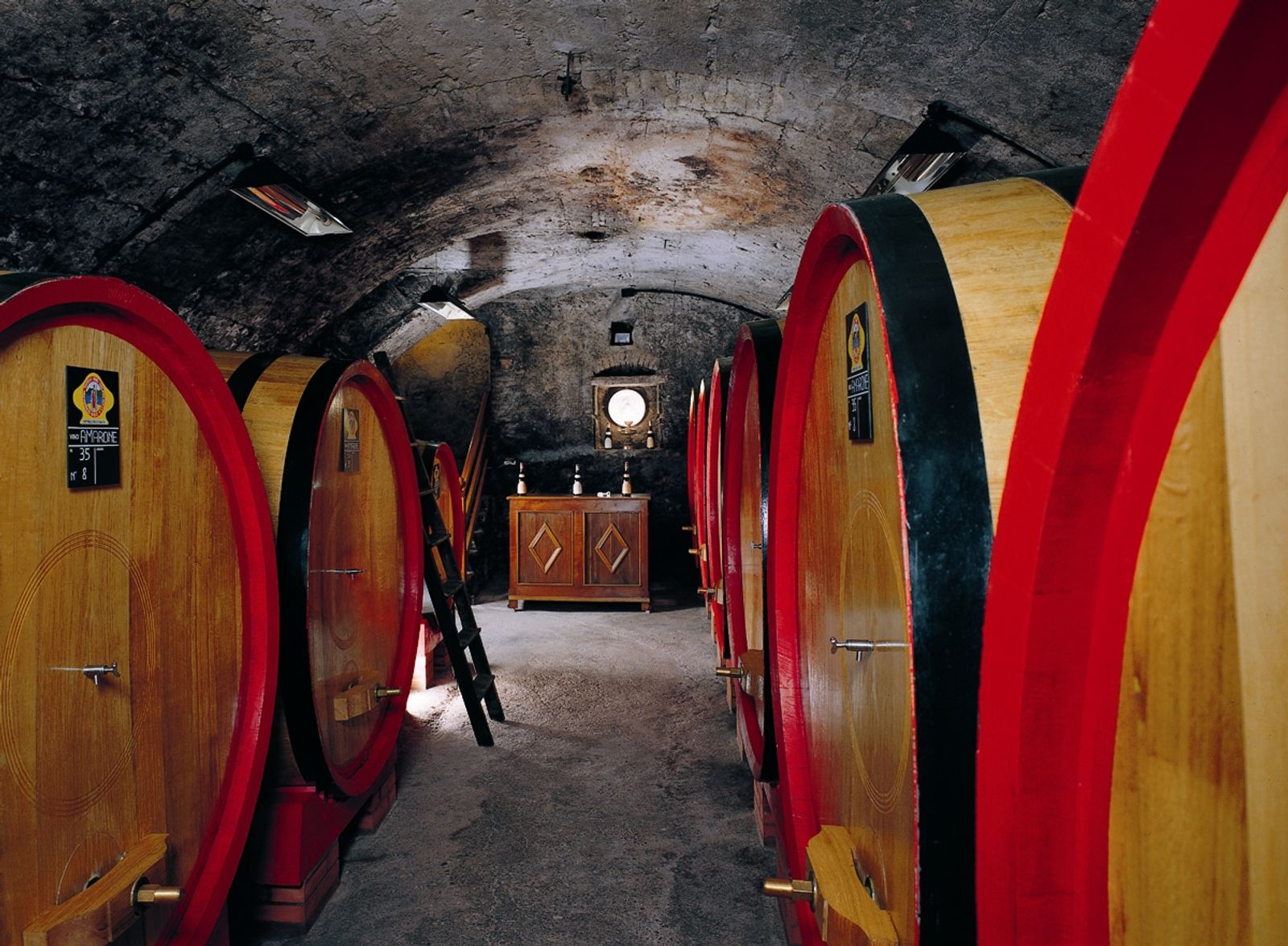 Grandi botti di rovere con cerchi rossi in una cantina con volta in mattoni e lanterne in vetro verde.