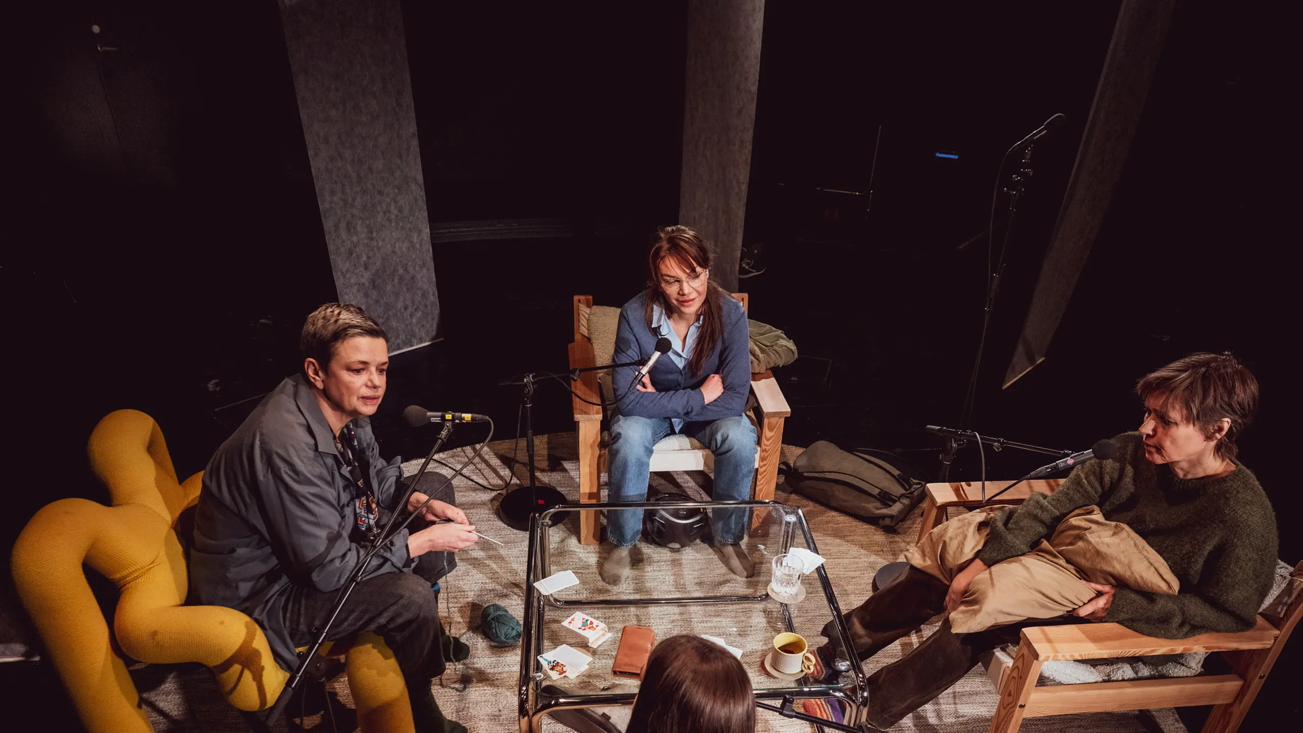 Fire personer sitter i ring rundt et bord med mikrofoner, kopper og papirer, i en scenisk samtalesetting.