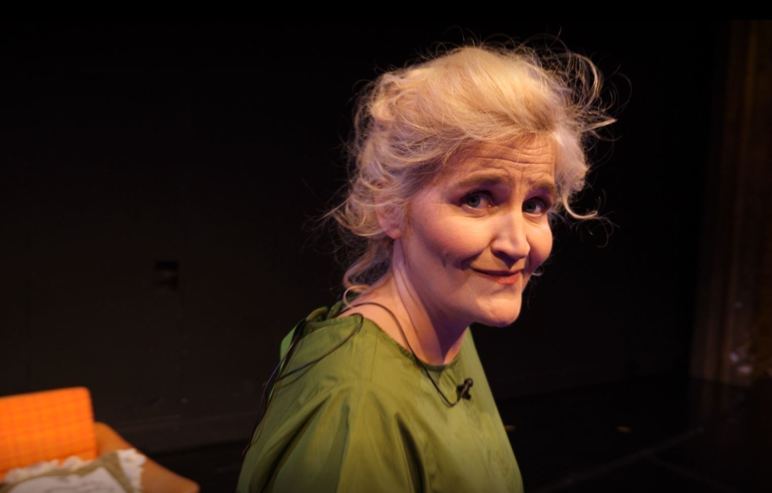 Smilende kvinne foran oransje sofa på mørk scene