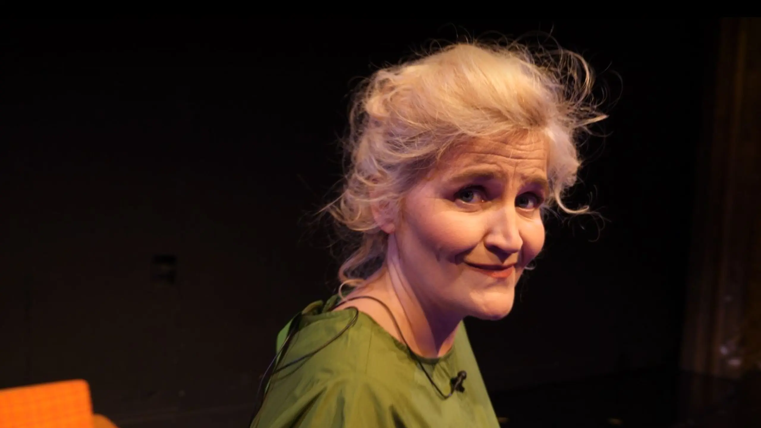 Smilende kvinne foran oransje sofa på mørk scene