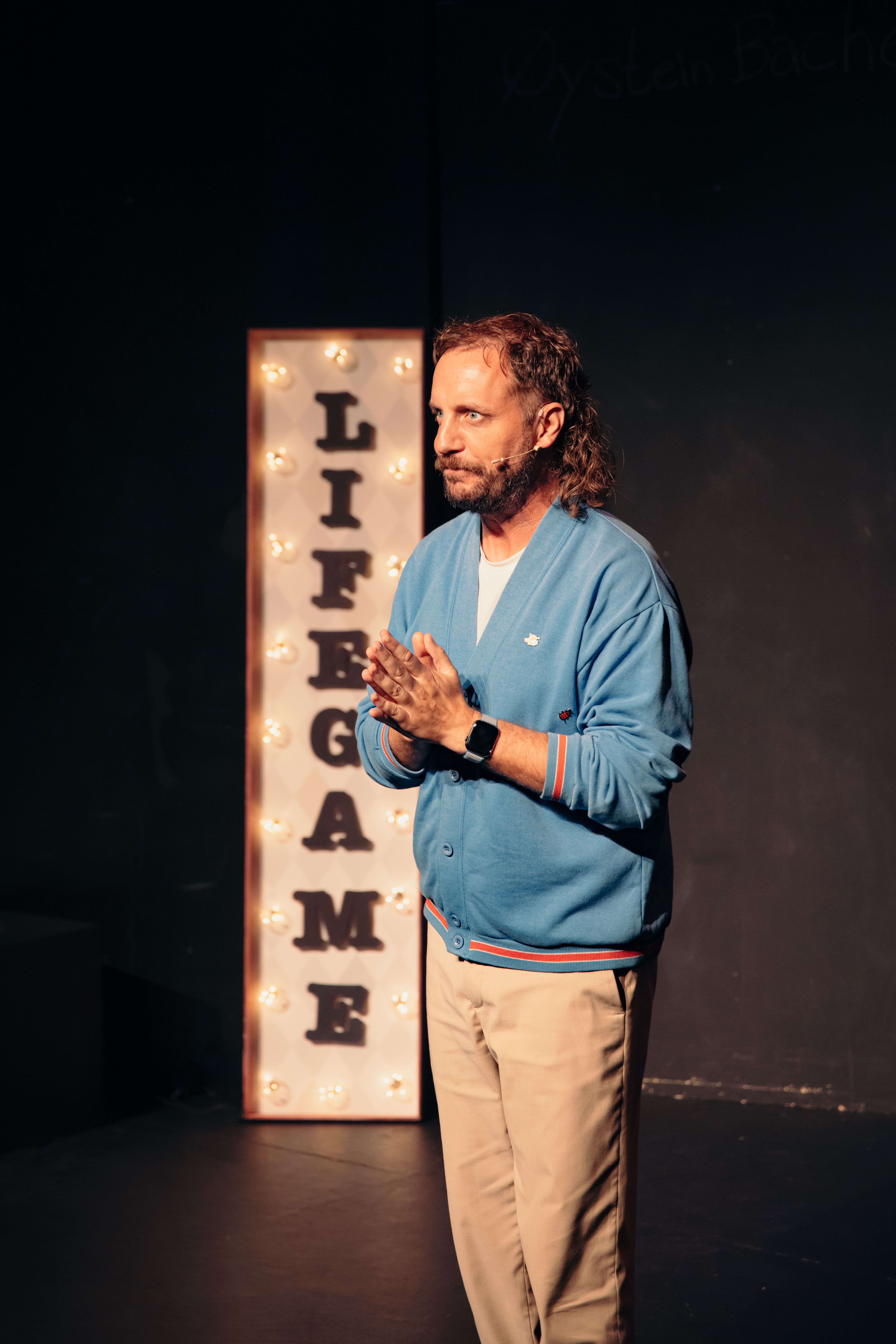 Person i blå cardigan står foran lysende skilt med teksten LIFE GAME og holder hendene samlet