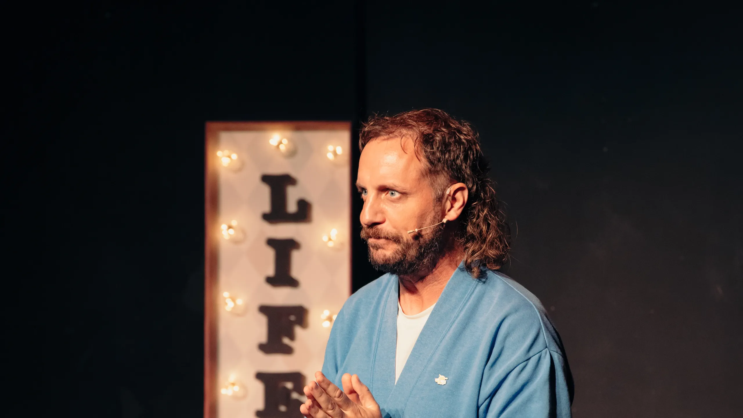 Person i blå cardigan står foran lysende skilt med teksten LIFE GAME og holder hendene samlet