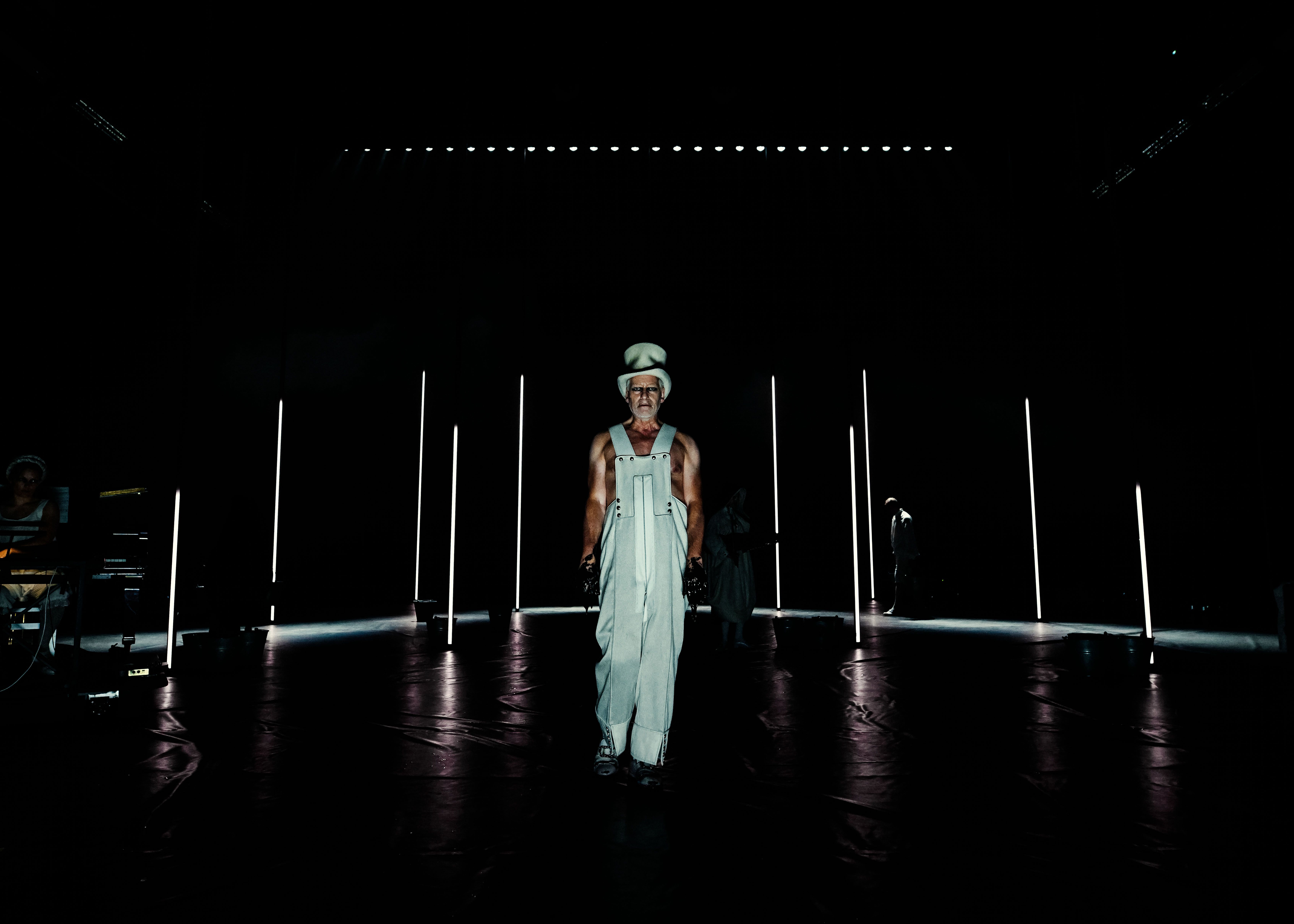 En mann i bunadsinspirert hvitt kostyme og hatt går sakte mot kamera på en mørk scene med lyskjegler. Han har tilgriset hendene i svart klissete væske