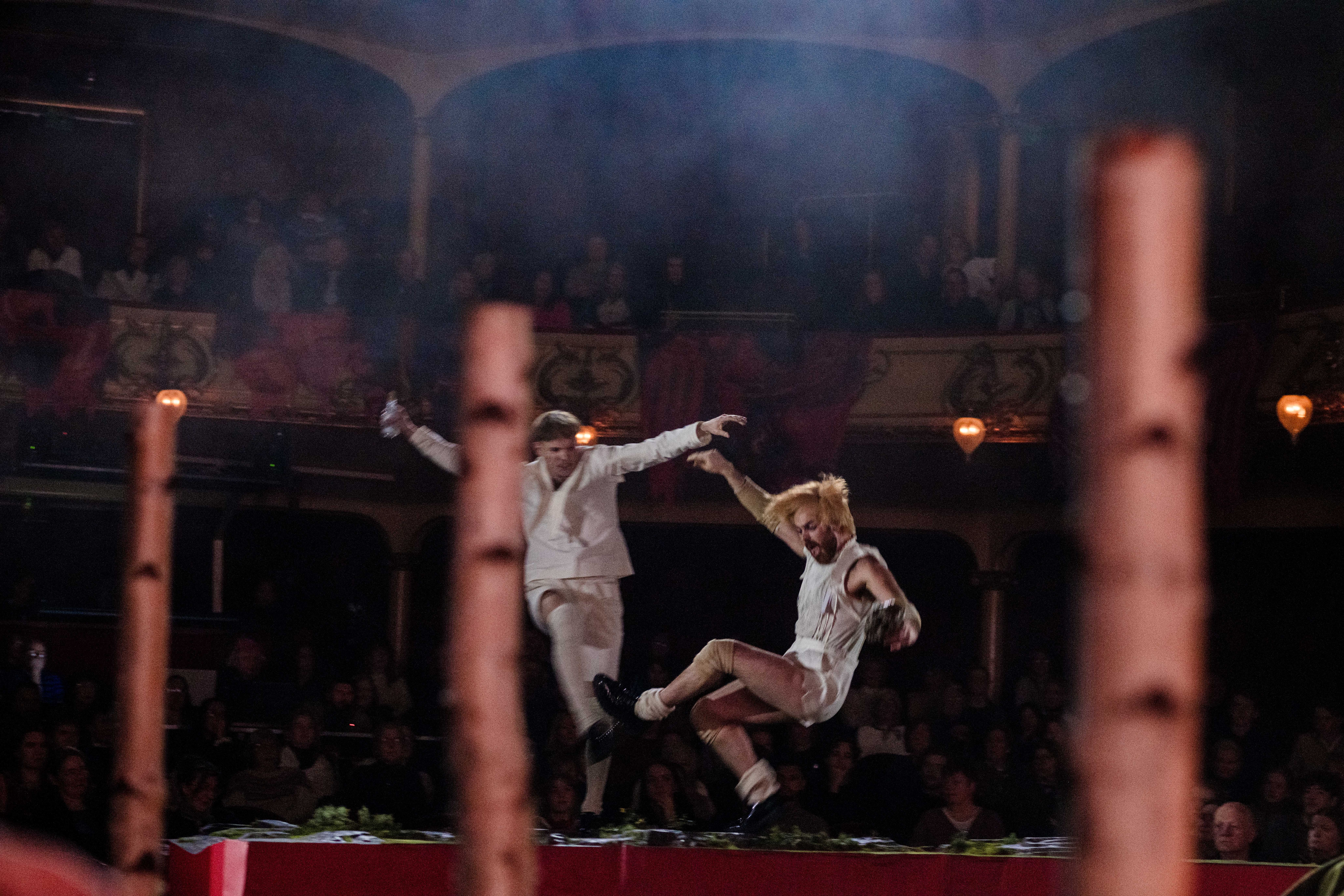 To scenekunstnere i hvitt hopper i luften på scenen, med uklare bjørkestammer i forgrunnen og publikum i bakgrunnen.
