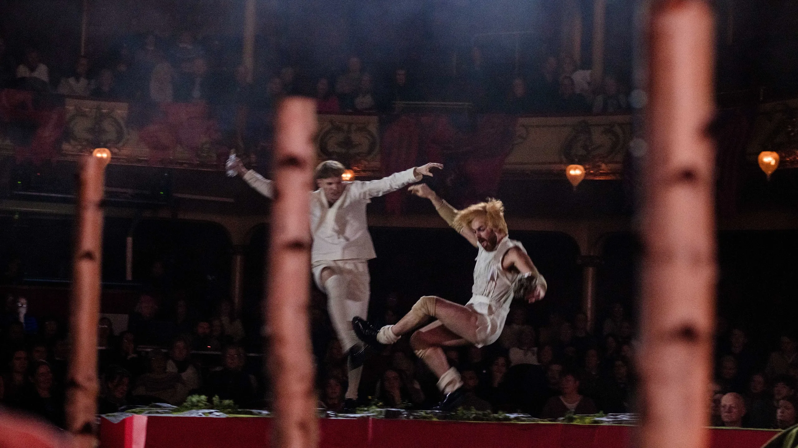 To scenekunstnere i hvitt hopper i luften på scenen, med uklare bjørkestammer i forgrunnen og publikum i bakgrunnen.