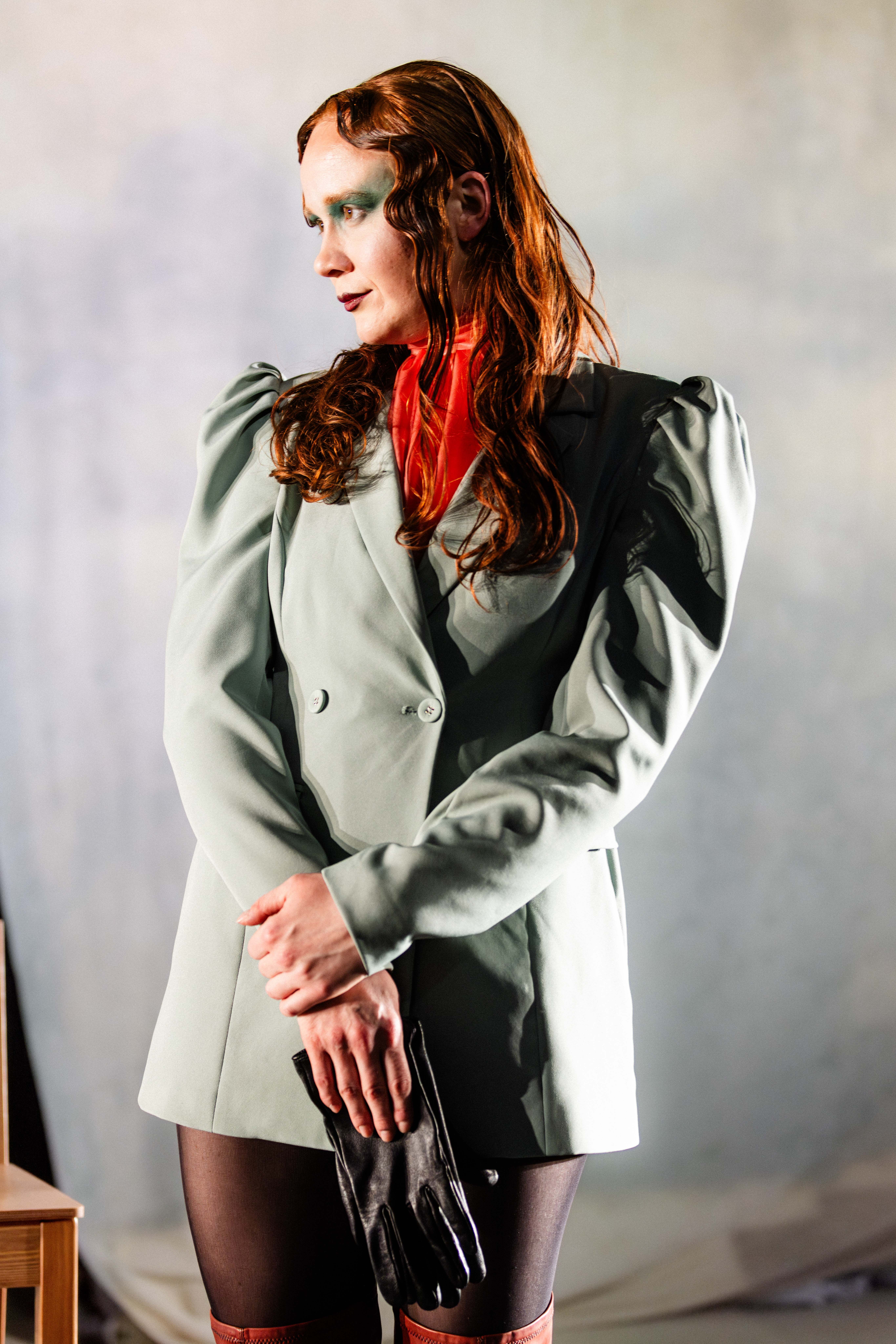 Person med langt rødt hår i grønn jakke og røde støvler står i scenelys og holder svarte hansker