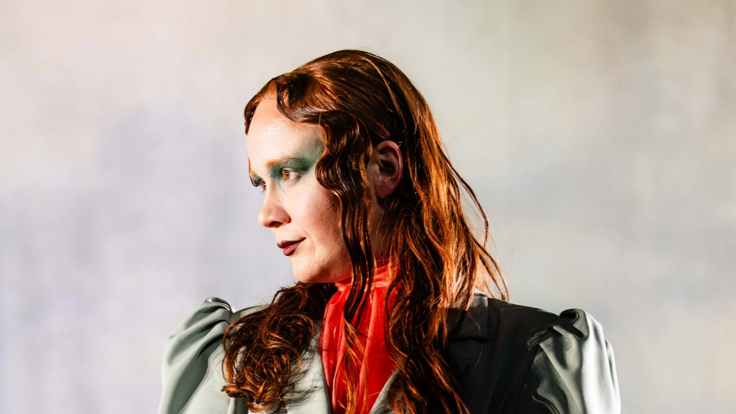 Person med langt rødt hår i grønn jakke og røde støvler står i scenelys og holder svarte hansker