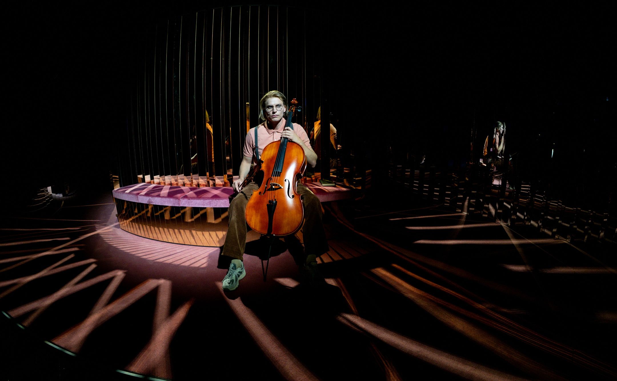 Person i rosa skjorte og brune bukser sitter i spotlight med en cello i fanget foran speil og mørk scenografi.