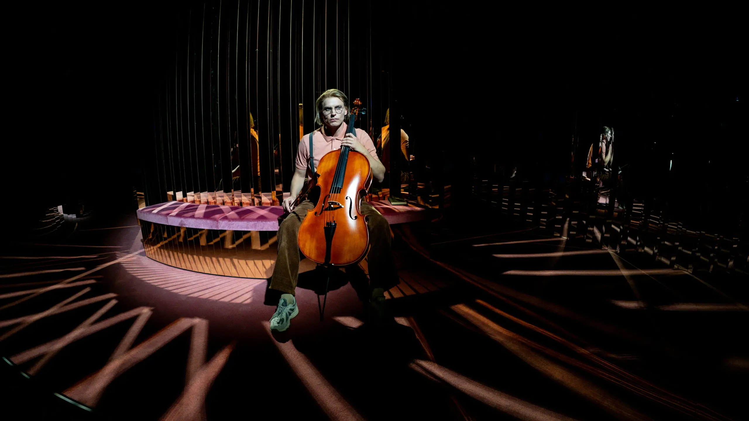 Person i rosa skjorte og brune bukser sitter i spotlight med en cello i fanget foran speil og mørk scenografi.