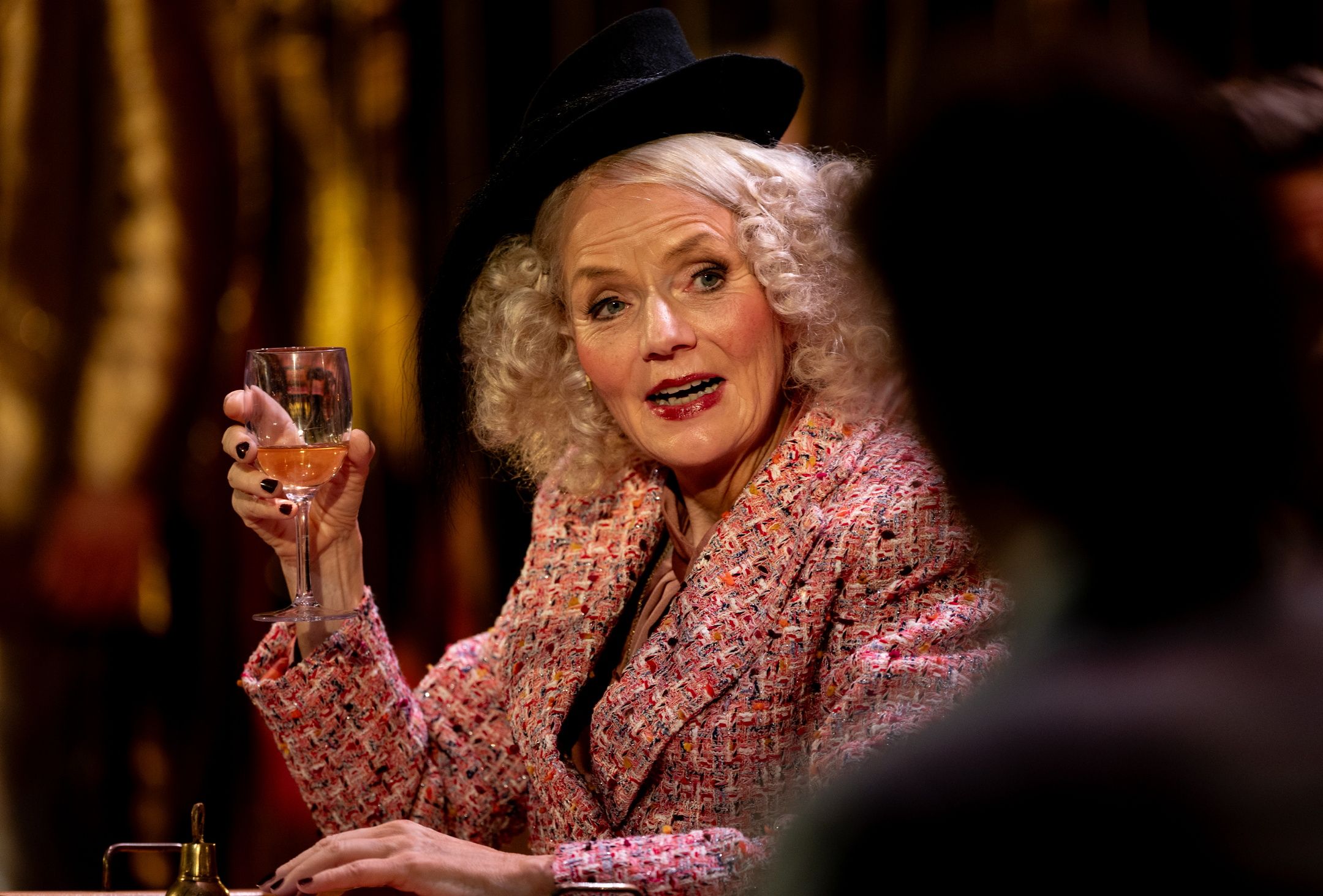 kvinne med lyst krøllete hår, hatt og fargerik jakke holder et vinglass og snakker engasjert.