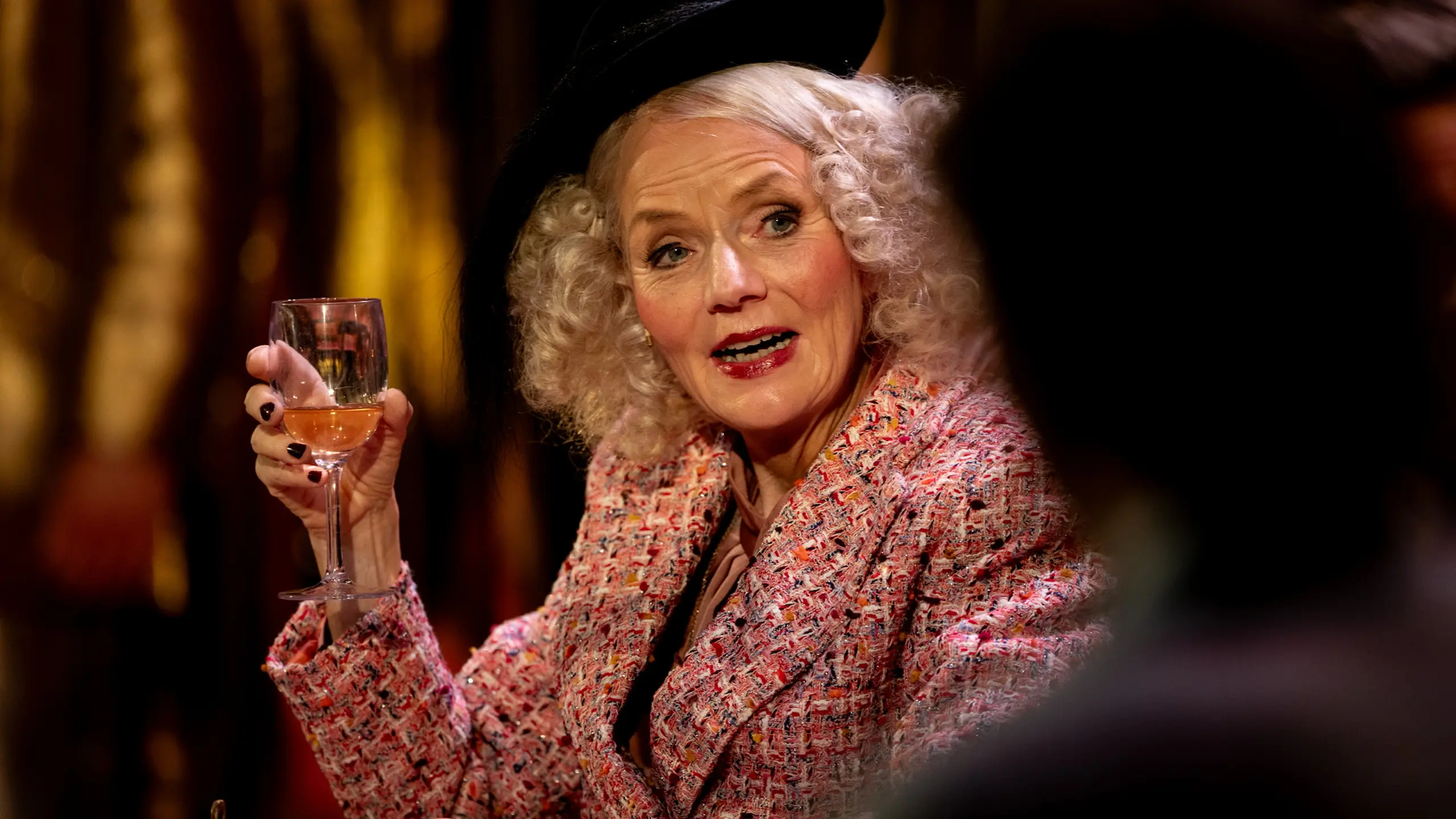 kvinne med lyst krøllete hår, hatt og fargerik jakke holder et vinglass og snakker engasjert.