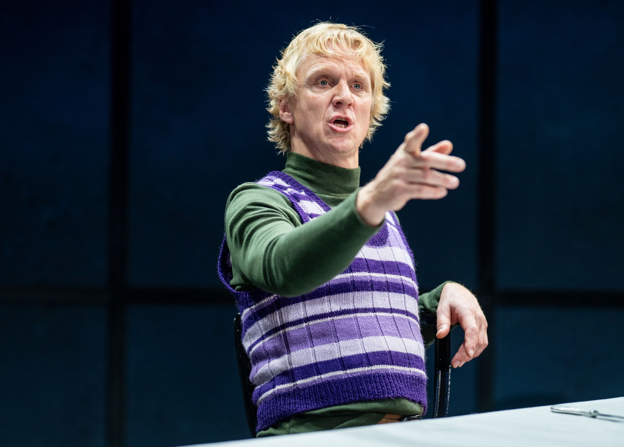 Nærbilde av mann i lilla og hvit stripete vest som peker mens han snakker.