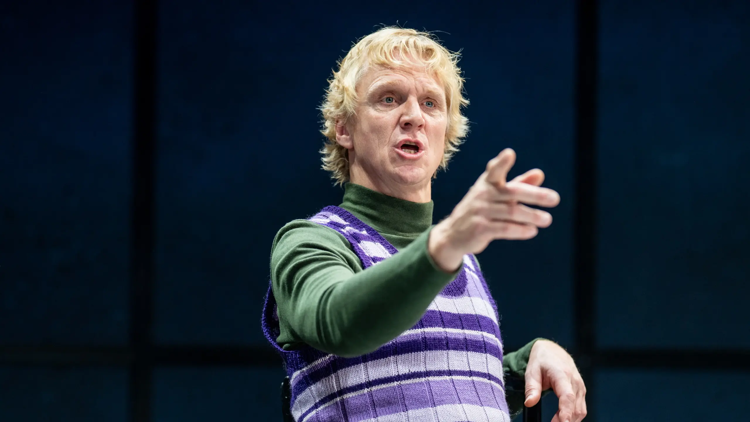 Nærbilde av mann i lilla og hvit stripete vest som peker mens han snakker.