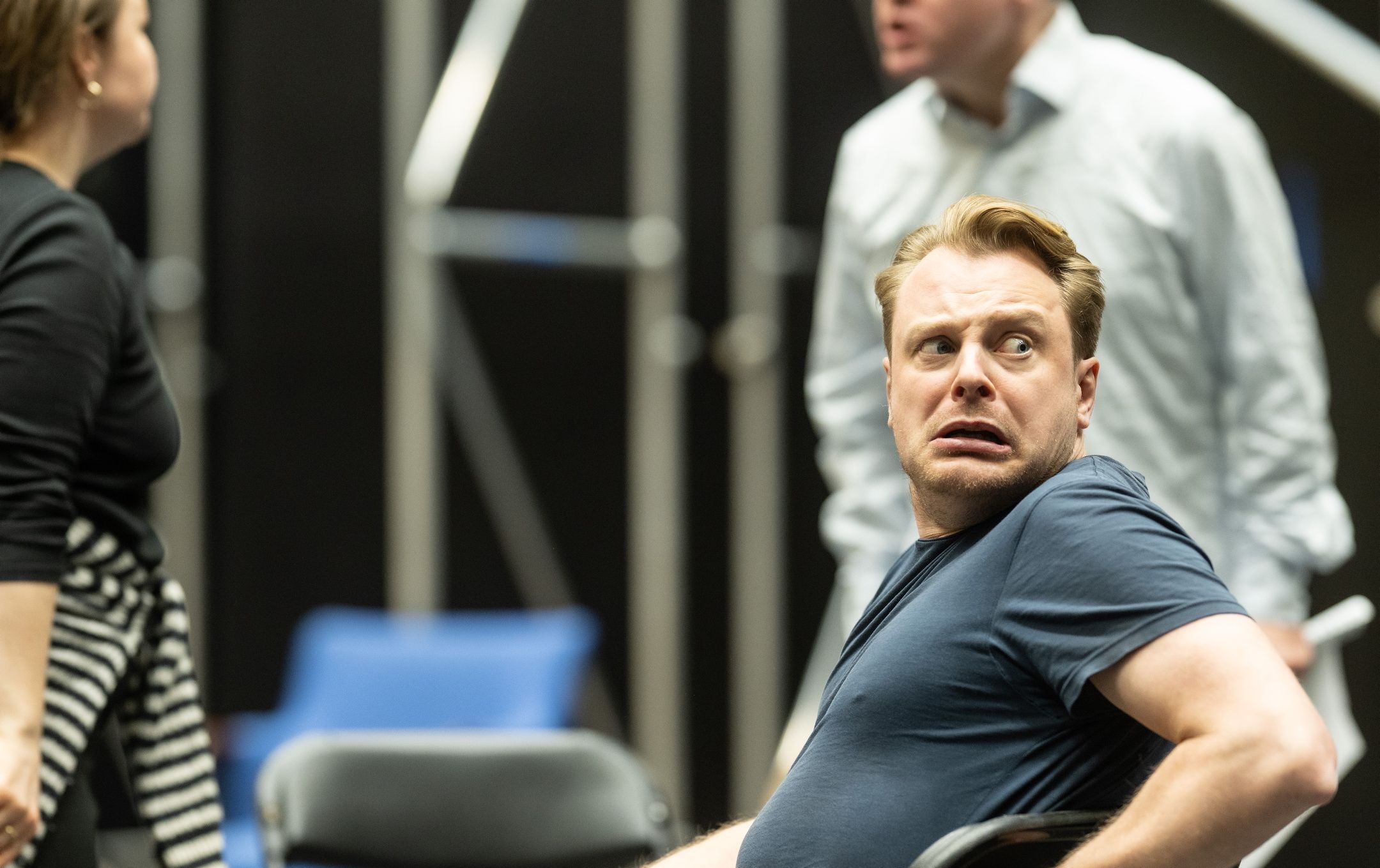 En mann i blå t-skjorte i forgrunnen ser skremt bakover mot noe utenfor bildet, mens to andre skuespillere er delvis synlige.