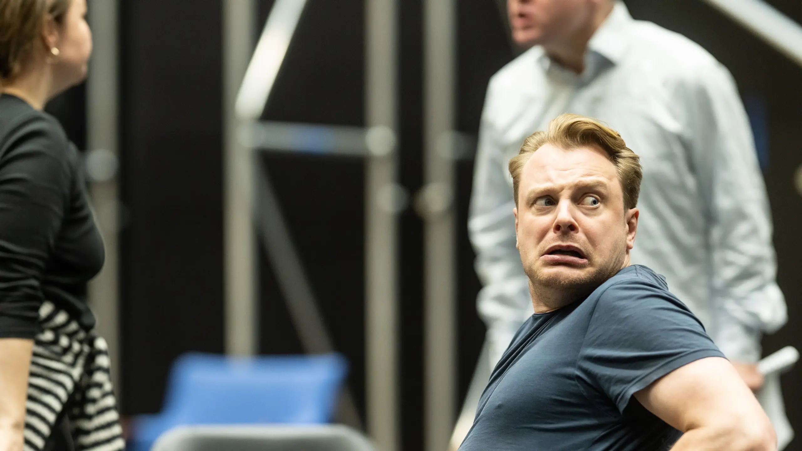 En mann i blå t-skjorte i forgrunnen ser skremt bakover mot noe utenfor bildet, mens to andre skuespillere er delvis synlige.
