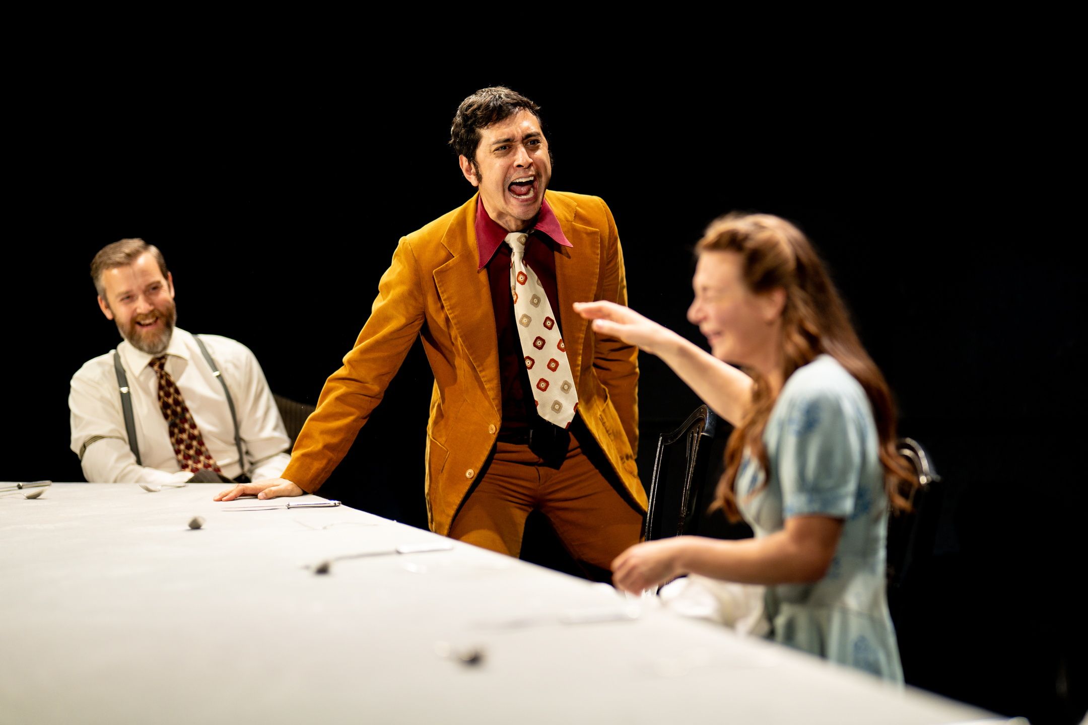 Tre personer rundt et bord, en mann i sennepsgul dress roper mens en kvinne peker og ler.