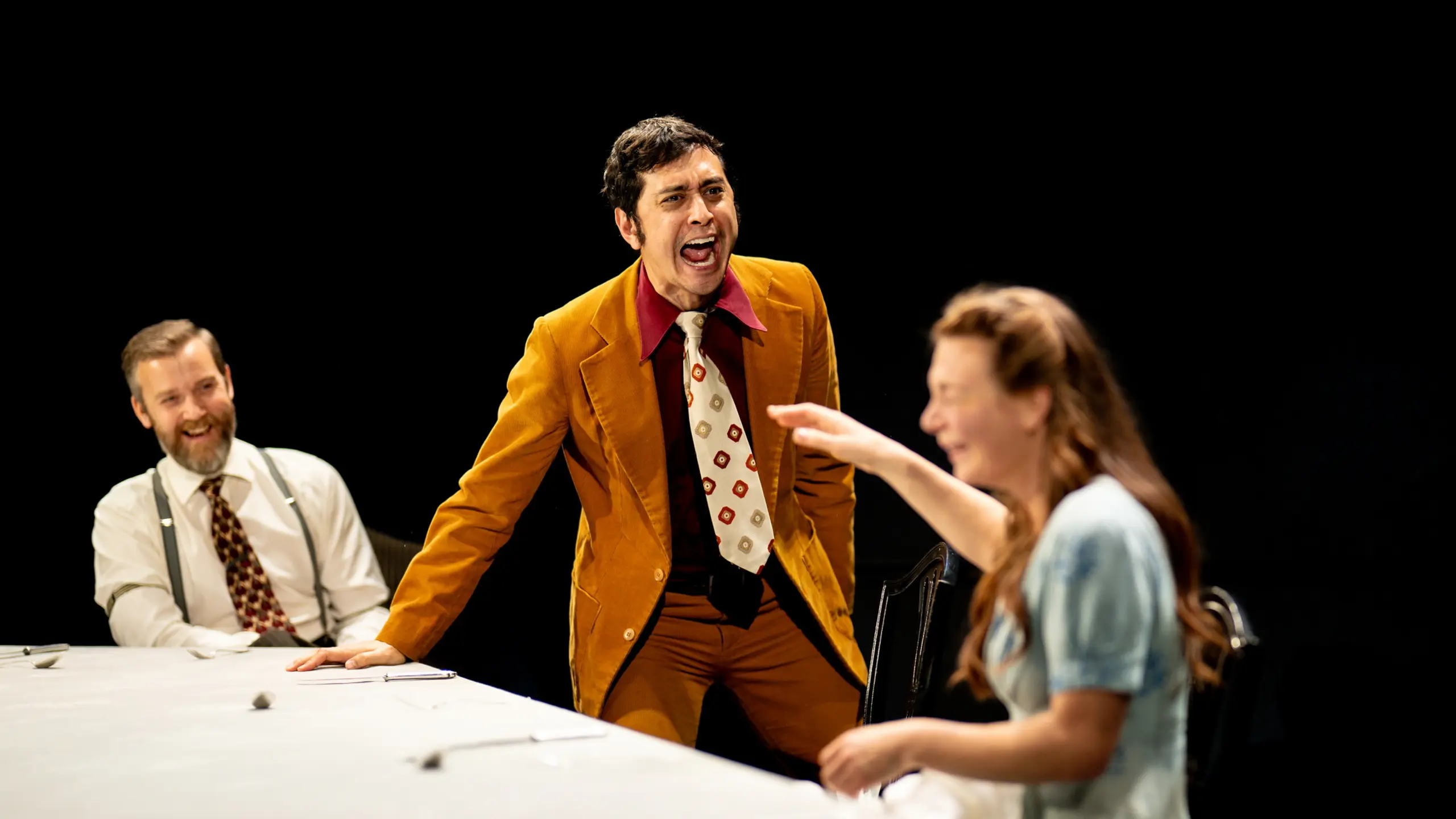 Tre personer rundt et bord, en mann i sennepsgul dress roper mens en kvinne peker og ler.