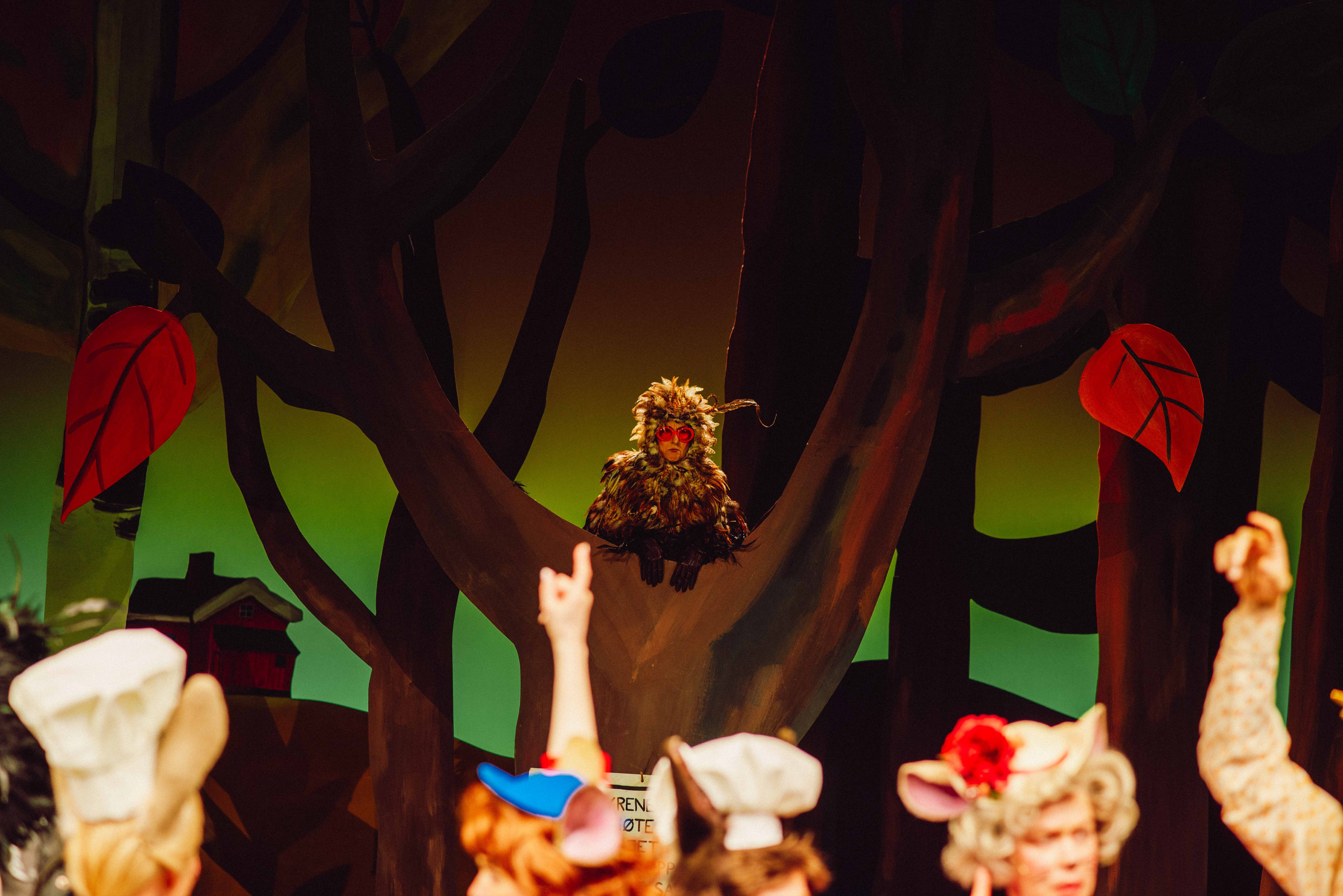 En person i ugledrakt med røde briller sitter i et tre, mens personer i forgrunnen myldrer rundt og peker