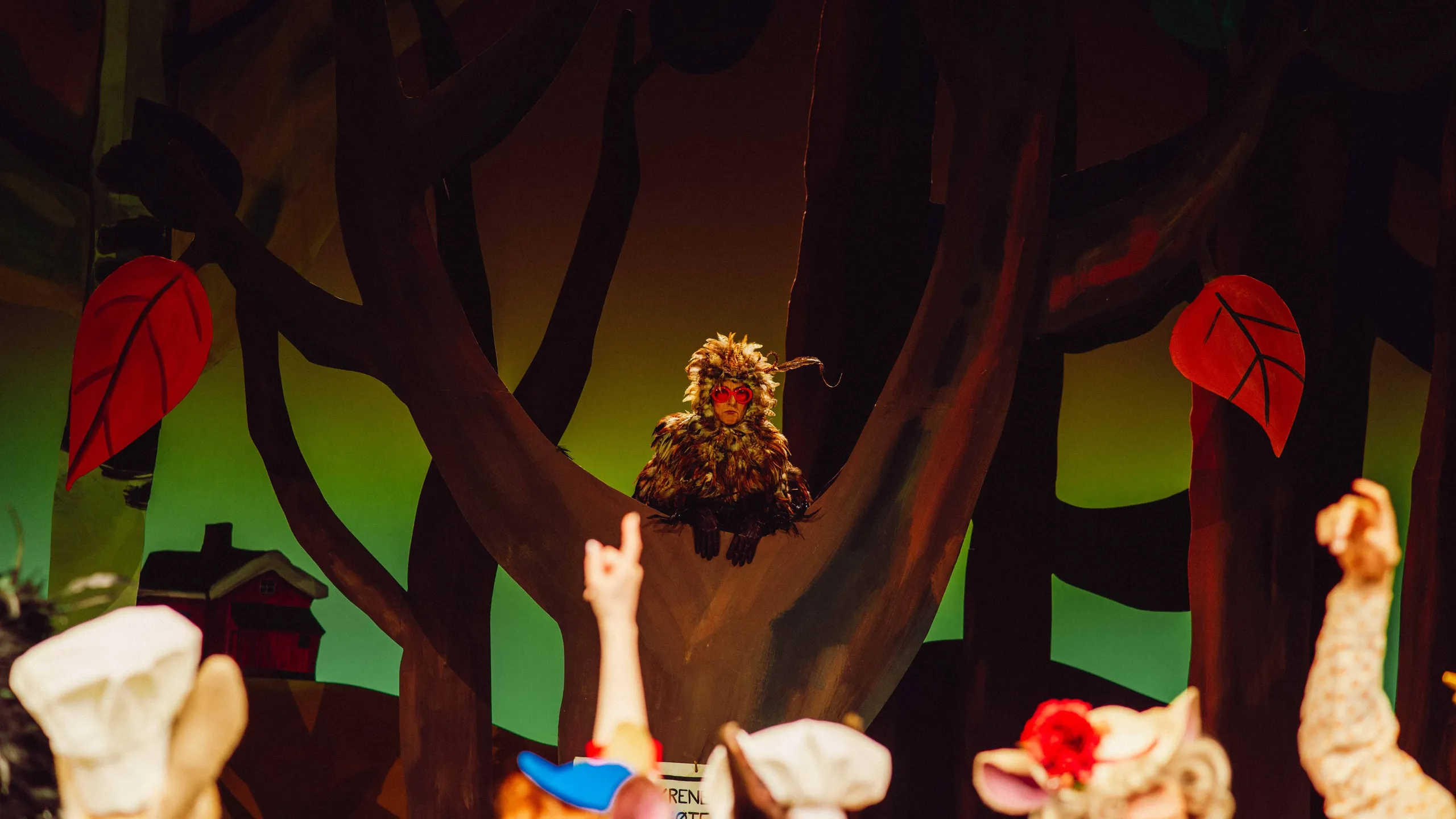 En person i ugledrakt med røde briller sitter i et tre, mens personer i forgrunnen myldrer rundt og peker