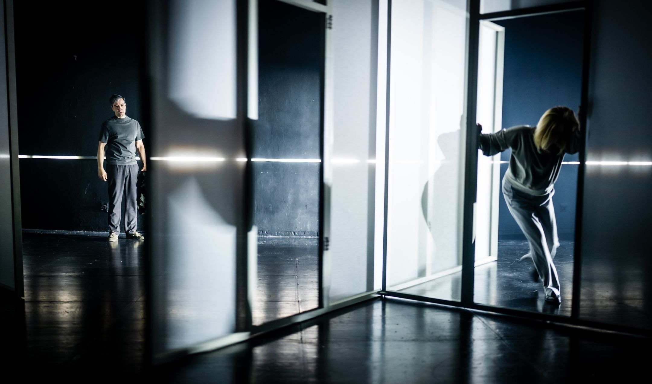 En kvinne i grå klær lener seg frem gjennom en glassdør, mens en mann står i bakgrunnen og ser på i et mørkt rom med stramt scenelys.