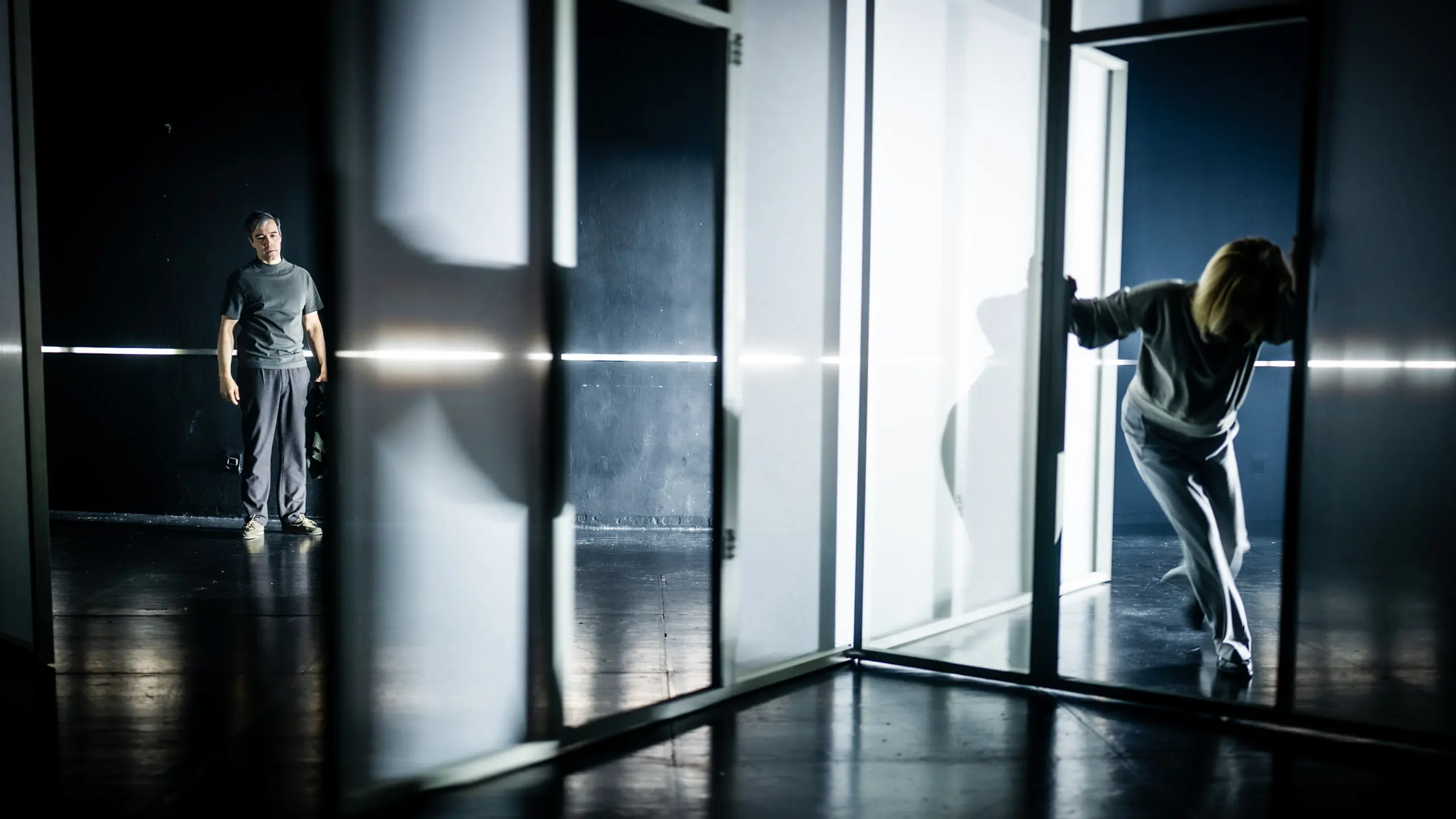 En kvinne i grå klær lener seg frem gjennom en glassdør, mens en mann står i bakgrunnen og ser på i et mørkt rom med stramt scenelys.
