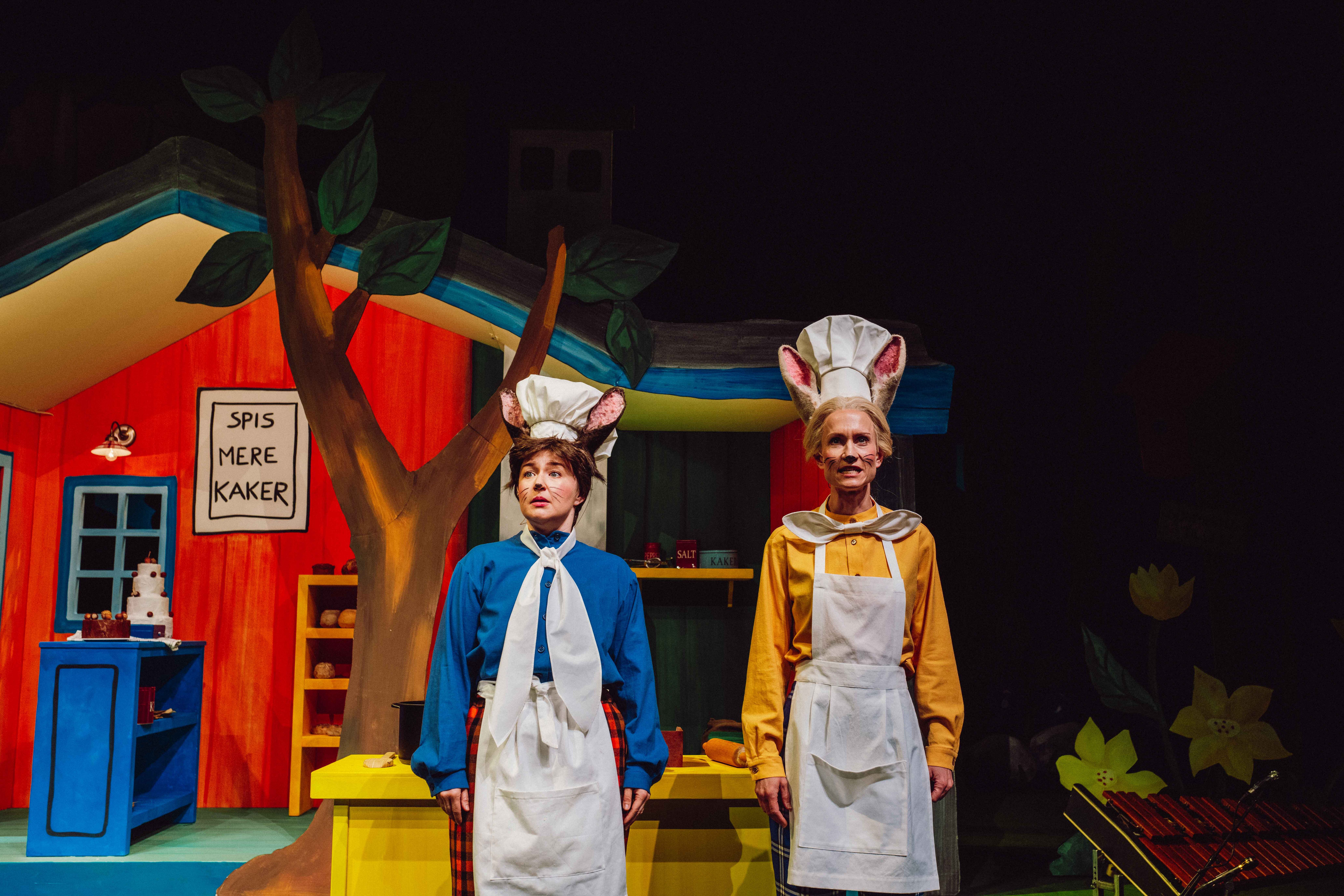 To bakere med museører står foran et fargerikt hus med skiltet 'Spis mere kaker'.